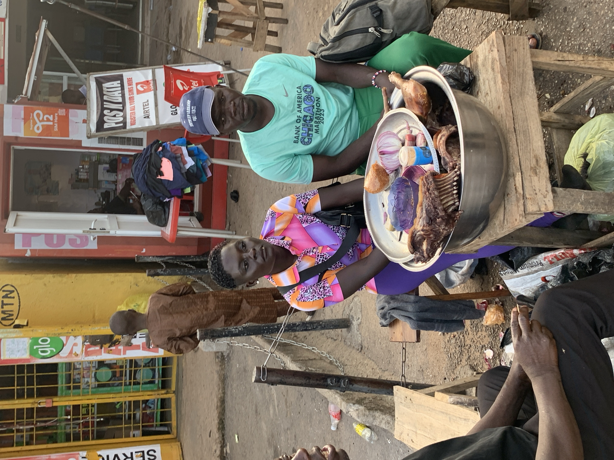 The image depicts a street scene in Jos, Nigeria. In the foreground, a woman in a vibrant, multi-colored shirt and purple leggings sits next to a man wearing a turquoise t-shirt with "BANK OF AMERICA CHICAGO MARATHON 2023" printed on it and green pants. They are seated on a wooden bench or stool. In front of them, on a wooden table, is a large metal bowl filled with cooked meat, including ribs and a leg of poultry. There are also red onions and a small purple dish containing what appears to be a dark sauce or condiment in the bowl. A black backpack is visible behind the man. To the left, another person's hands and lap are visible, holding a small stick. In the background, a young boy wearing a brown tunic stands near a yellow building with various signs, including "MTN" and "Glo." A red table with folded clothes is also visible. The environment appears to be a busy street with pavement and some scattered debris.
