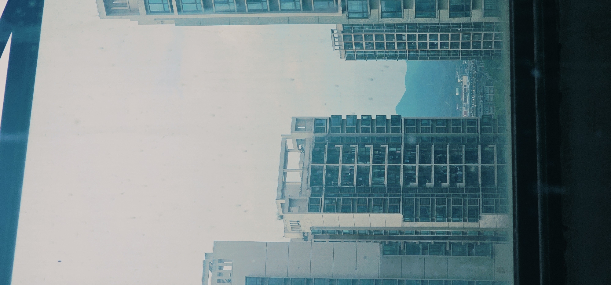 The image captures a view of the Chongqing High-tech Zone in China, seen through a slightly dirty window. Multiple modern high-rise apartment buildings dominate the scene, characterized by their rectangular structures and numerous windows. The architecture suggests a dense urban environment. In the background, a hazy, blue mountain range is visible, indicating a hilly or mountainous terrain surrounding the city. The sky is overcast and pale, contributing to a somewhat muted and cool color palette with dominant blues and grays. The overall impression is one of a contemporary cityscape, possibly captured during daytime under cloudy conditions.