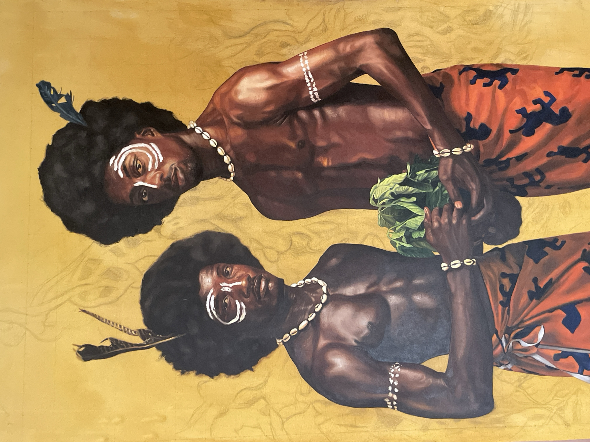 Two dark-skinned men lie horizontally on a gold background. Each man has large afro hair adorned with a single feather. White paint marks encircle their eyes. Both men wear a necklace of white shells and a matching bracelet. The men are shirtless and wear orange fabric draped around their waists, patterned with dark blue silhouettes of a figure. The upper man holds a small bundle of green leaves. The location is Ado, Nigeria.