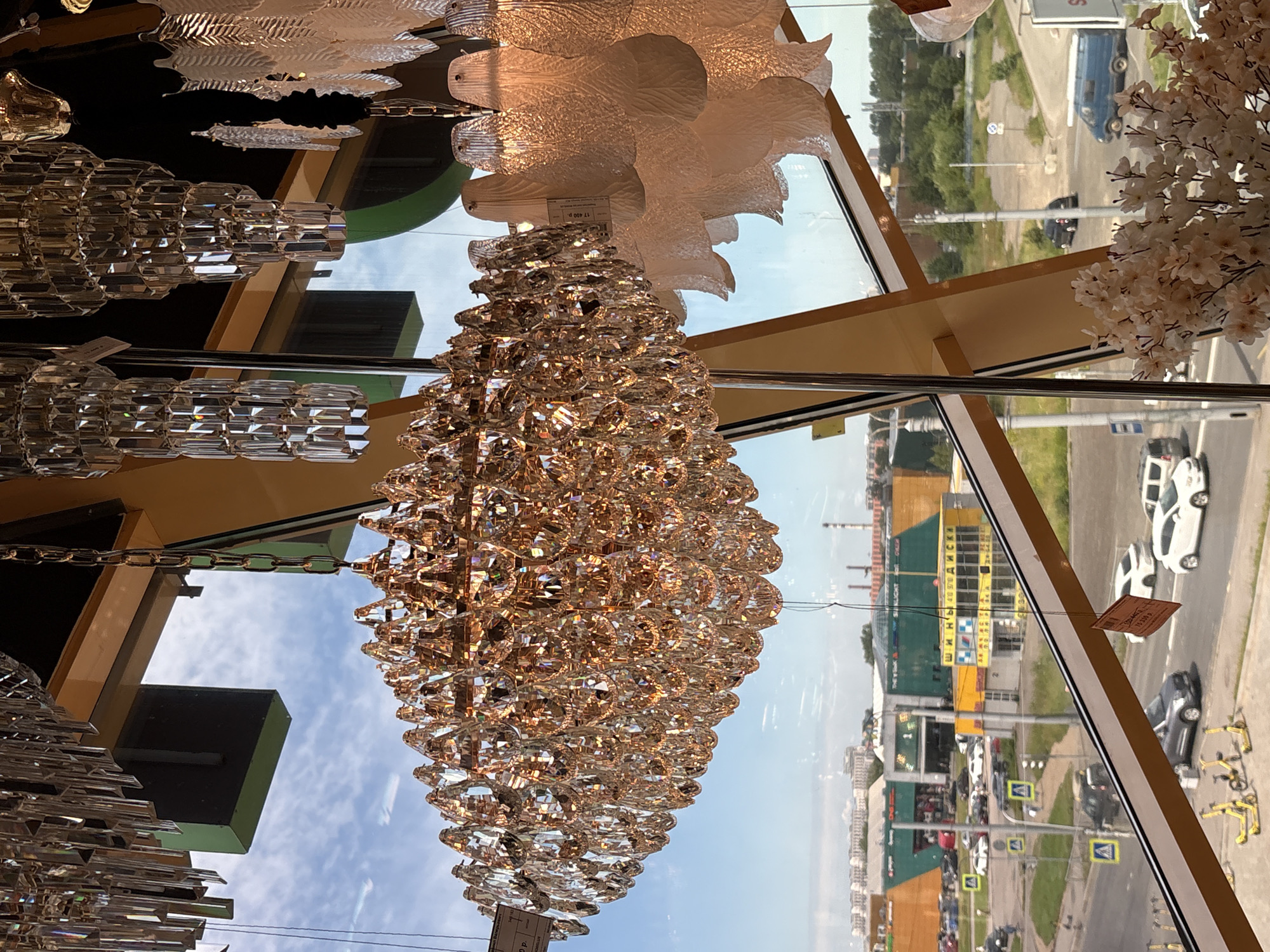 The image was taken on **June 15, 2024**, at **1:30 PM** from inside a building with large windows, looking out at a commercial area in **Saint Petersburg, Russia**. The view is dominated by several ornate chandeliers, one large and golden, and others made of clear faceted crystals, hanging in the foreground. Through the windows, a sunlit street scene is visible with cars, a blue van, yellow electric scooters, and several retail buildings. One building has a prominent yellow sign with Russian text. The sky is blue with scattered clouds. The photo appears to be taken from a store or showroom displaying luxury lighting fixtures. There are no visible people in the scene.