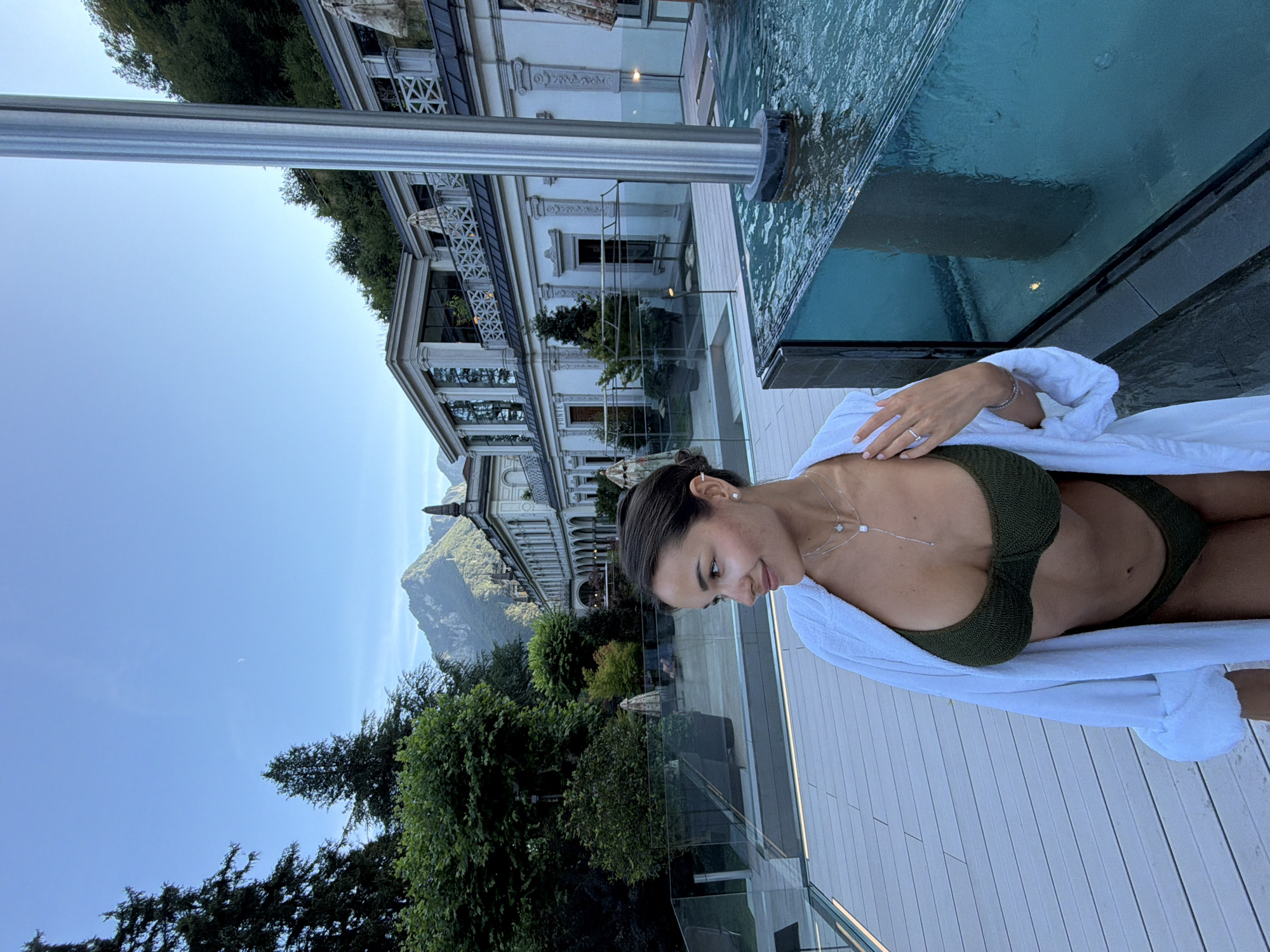 A woman in a dark green bikini and a white robe stands on a wooden deck next to a glass-enclosed pool. The woman is looking to her left, with her right hand resting on her hip. Behind her, a large white building with ornate architectural details is visible, nestled against a lush green hillside. Trees are scattered throughout the landscape, and mountains rise in the background under a clear blue sky with a crescent moon. The scene appears to be at a resort or spa in San Pellegrino Terme, Italy.