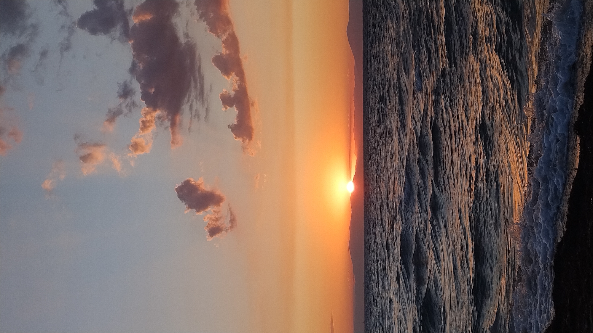This image depicts a serene sunset over the Aegean Sea, viewed from the coast of Neoi Epivates, Greece. The sun is partially obscured by a low-lying mountain range on the horizon, casting a warm orange glow across the sky and reflecting on the rippling water. Wispy clouds, tinged with pink and purple, are scattered across the pale blue and orange sky. Gentle waves are breaking on a dark, pebbly shore in the foreground, creating white foam. No people or specific structures are visible, emphasizing the natural beauty of the coastal landscape at dusk.