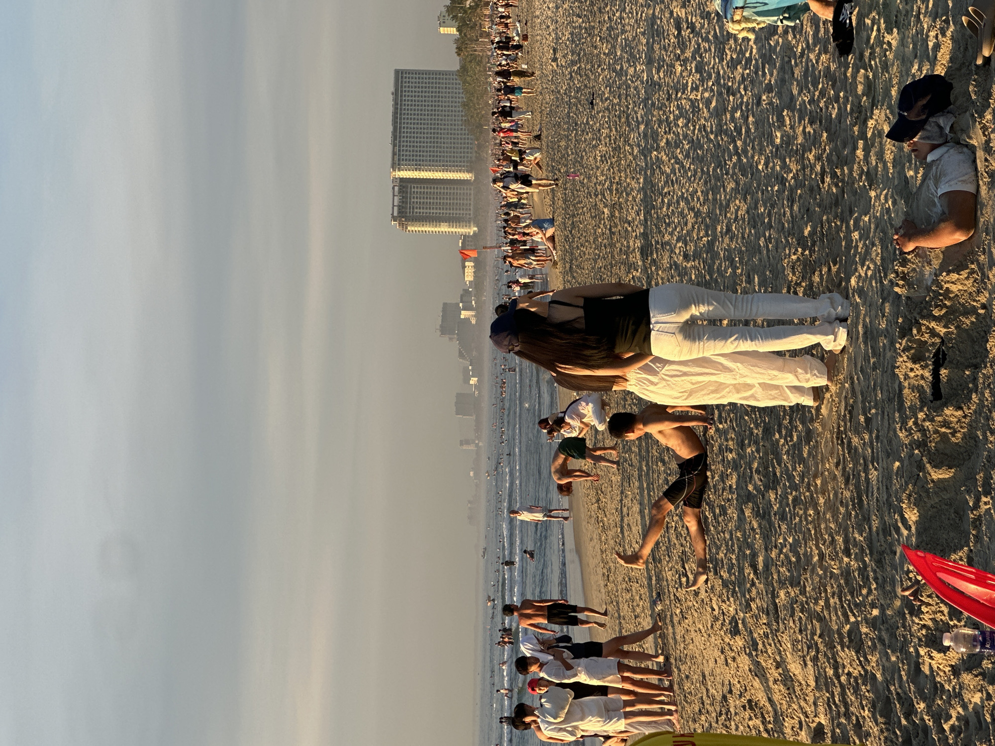 This image depicts a crowded beach scene at sunset in Đà Nẵng, Vietnam. In the foreground, several people are seen on the sandy beach. A man lies on his back in the sand, with his hands clasped above his head. To his left, a red object, possibly a boogie board, lies partially buried in the sand next to a water bottle. In the mid-ground, a group of people stand and walk along the shoreline. Further out, the ocean is filled with swimmers and small boats. In the background, a modern, large hotel complex with multiple stories and balconies is visible, as are other buildings suggesting an urban environment beyond the beach. The sky is hazy with the soft light of the setting sun.