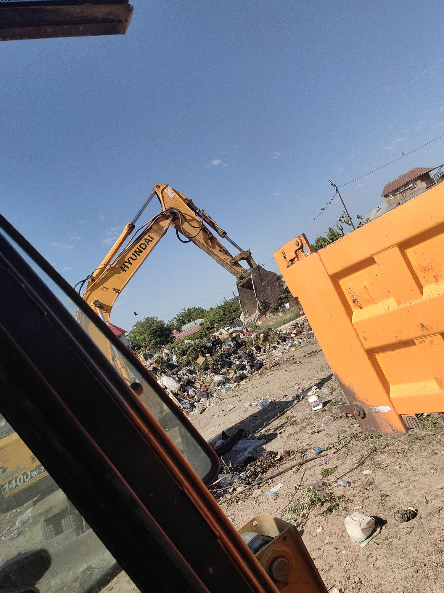 This image depicts a construction site or landfill in the city of Shymkent, Kazakhstan. In the foreground, the metallic structure of an orange dump truck and the yellow arm of a Hyundai excavator are visible. The excavator's bucket is positioned over a large pile of refuse, suggesting an ongoing waste disposal operation. The ground is uneven and littered with debris. In the background, a residential building with a tiled roof and trees can be seen against a clear blue sky. The overall scene suggests a dusty, outdoor environment with evidence of construction or waste management activities.