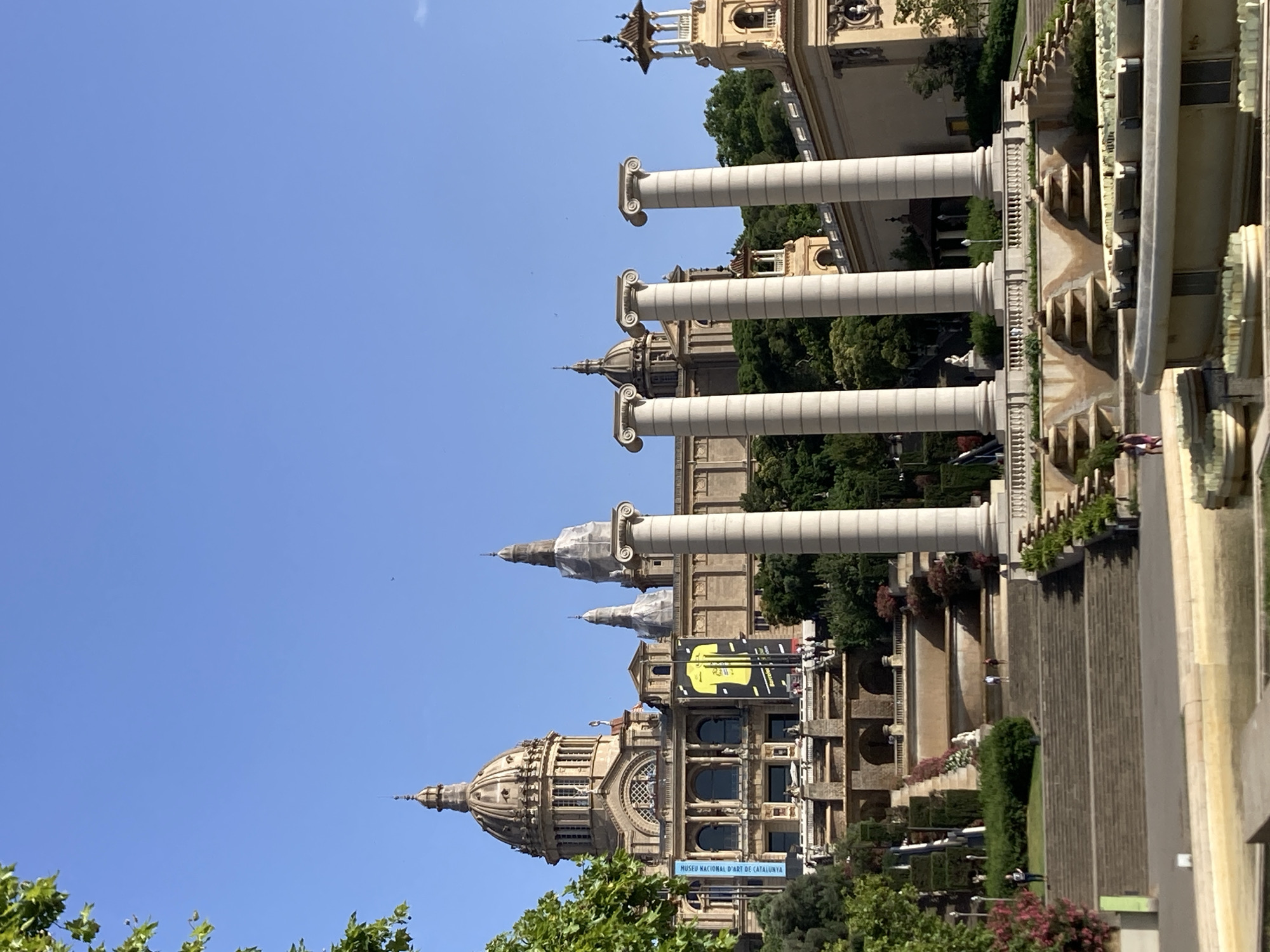 The image depicts the Palau Nacional, the National Palace of Catalonia, located on Montjuïc hill in Barcelona, Spain. The majestic building, characterized by its large central dome and neoclassical architecture, is set against a clear blue sky. In the foreground, a series of grand Ionic columns stand tall, framing a tiered fountain and cascading stairways adorned with lush greenery and flowering bushes. Several people can be observed on the steps and pathways, enjoying the scenic surroundings. The scene is bathed in bright sunlight, indicating a clear and pleasant day.