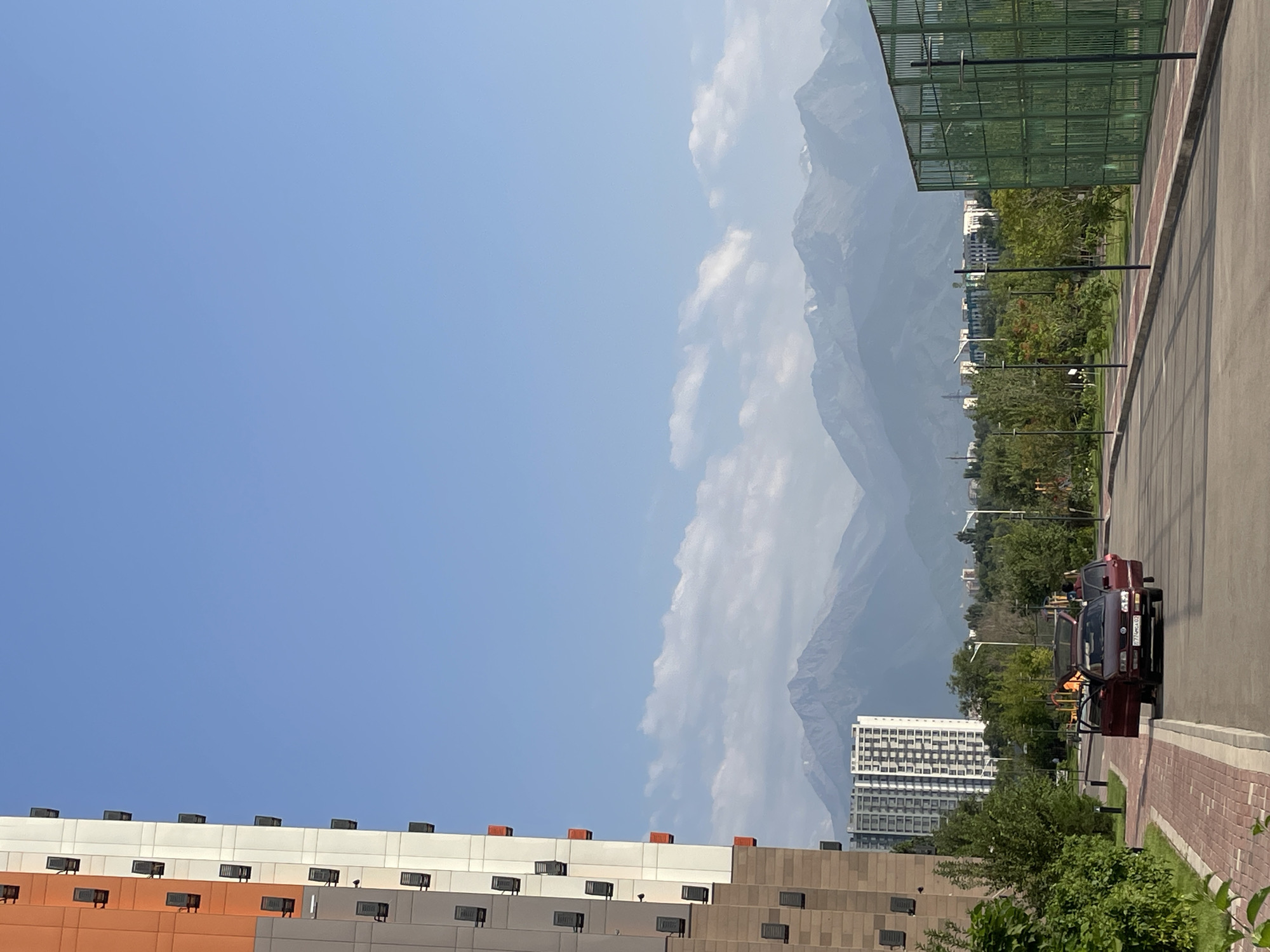 An exterior, daytime, eye-level, vertical shot shows a street in Almaty, Kazakhstan, with a modern apartment building on the left and mountains in the background. The building on the left is tall and modern, with a façade featuring sections of white, orange, and tan. Numerous dark rectangular window units are visible. In the middle ground, a street runs horizontally across the image. Two red cars are parked on the right side of the street, their doors open, with at least one person visible inside one of them. Beyond the street, there is a line of trees and other buildings, including a tall, white apartment building with a grid-like pattern of windows. In the background, majestic mountains with snow-capped peaks rise against a clear blue sky with scattered white clouds. A green metal fence structure is visible on the right side of the image. The scene suggests a residential area on the outskirts of the city, close to the mountains.