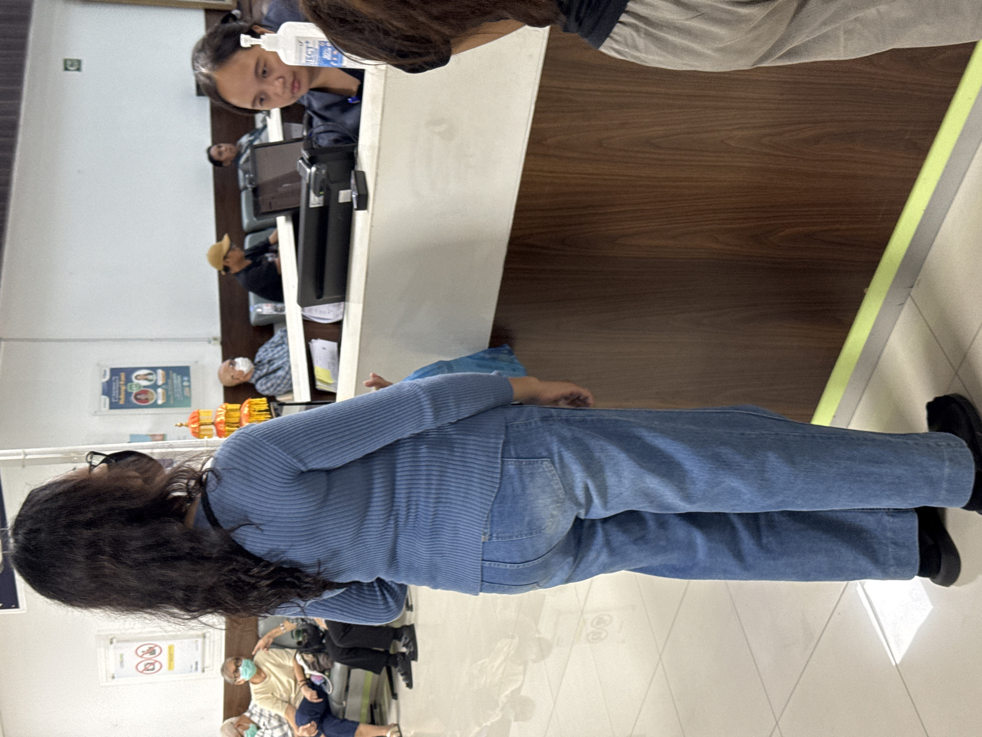 A medium shot captures a scene inside what appears to be a public service counter, possibly a hospital or government office, in Kuta, Indonesia. The primary subject is a woman from behind, wearing a blue knitted sweater and light blue denim jeans. She has long, dark, wavy hair and is wearing glasses and a black face mask. To her right, at a counter, a woman with dark hair tied back is interacting with another person, with a hand sanitizer bottle visible on the counter. In the background, several people are seated on a bench, some wearing face masks, suggesting adherence to health protocols. The counter is made of a light-colored material with a dark wood-patterned base. Posters are visible on the wall behind the seated individuals. The floor is tiled in a light color. The overall impression is of an organized, functional indoor space.