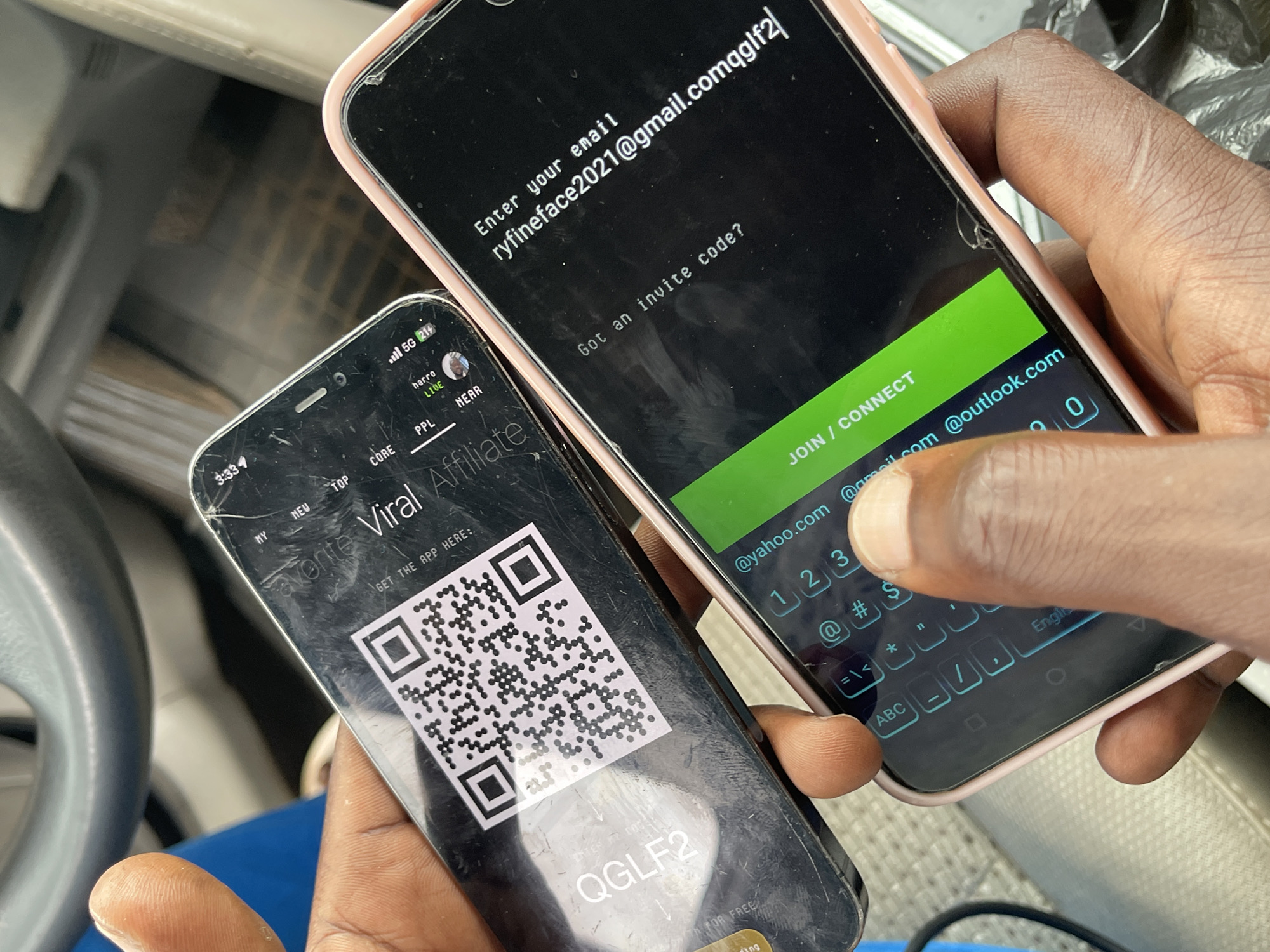 The image shows a close-up, overhead view of two smartphones being held by a person's hands. The phones are positioned diagonally across the frame, with the left phone being older and showing significant wear, and the right phone being newer with a pink case.
On the older, black smartphone on the left, a QR code is prominently displayed, suggesting a digital invitation or login. Above the QR code, the text "Viral Affiliate" is visible, along with smaller text that appears to be navigational elements or app features like "NY", "NEW", "TOP", "CORE", "PPL", "LIVE", "harro", "NEAR", and "5G". The phrase "GET THE APP HERE:" is also present. A code "QGLF2" is visible in the lower left portion of this phone's screen.
The newer, pink-cased smartphone on the right displays a login or signup screen. The text "Enter your email" is at the top, followed by an email address: "ryfnamefont2021@gmail.comqqglf2". Below this, there's a question: "Got an invite code?". A prominent green button with the text "JOIN / CONNECT" is at the bottom of the screen. A keyboard is partially visible, with keys displaying characters and numbers. The background of this phone's screen also shows partial email addresses like "@yahoo.com" and "@outlook.com".
The overall environment appears to be indoors, possibly a vehicle interior, given the blurred background elements that resemble car interiors. The lighting suggests daylight. The image is focused on the phones and hands, indicating the subject's attention is directed towards the screens and the task of entering information or connecting. The image does not provide any specific landmarks or elements that would place it definitively in Galadimawa Village, Nigeria.