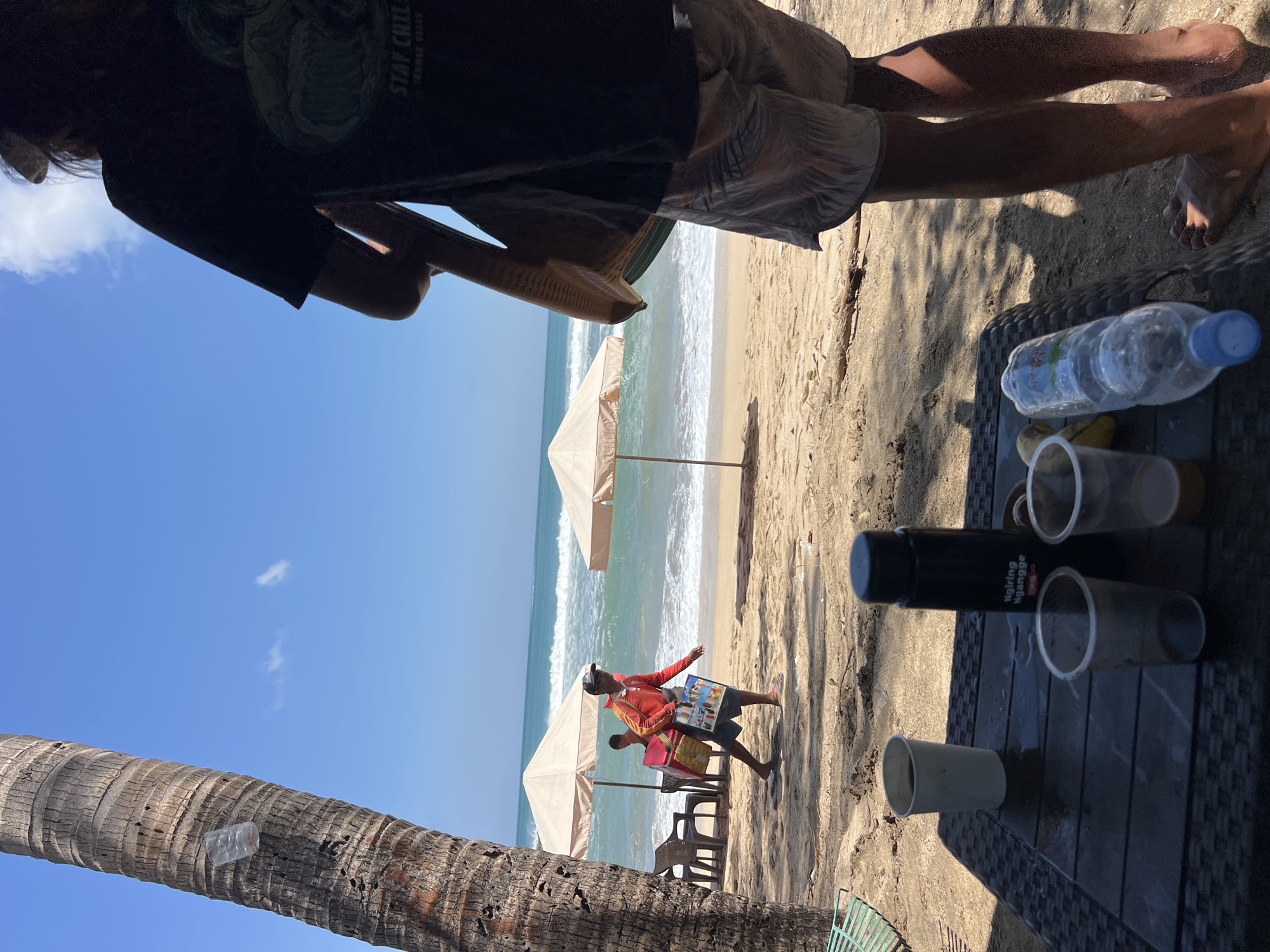 The image depicts a beach scene in Kuta, Indonesia, on a sunny day. In the foreground, a table made of woven material holds several items: a white paper cup with a dark liquid, a black thermos with text that appears to say "Ngiring Ngangge", and two clear plastic cups, one with some liquid in it. A partially visible bottle of water with a blue cap is also on the table, along with a banana. To the right, the lower legs and feet of a person wearing patterned shorts are visible. The background shows a sandy beach with a palm tree on the left. Further back, two beige beach umbrellas are set up, with chairs underneath one of them. A vendor in a red shirt and dark shorts, carrying a red cooler bag and a flat rectangular item, walks along the beach towards the ocean. The ocean has moderate waves under a bright blue sky with a few scattered clouds. The overall impression is a relaxed beach atmosphere with commercial activity.