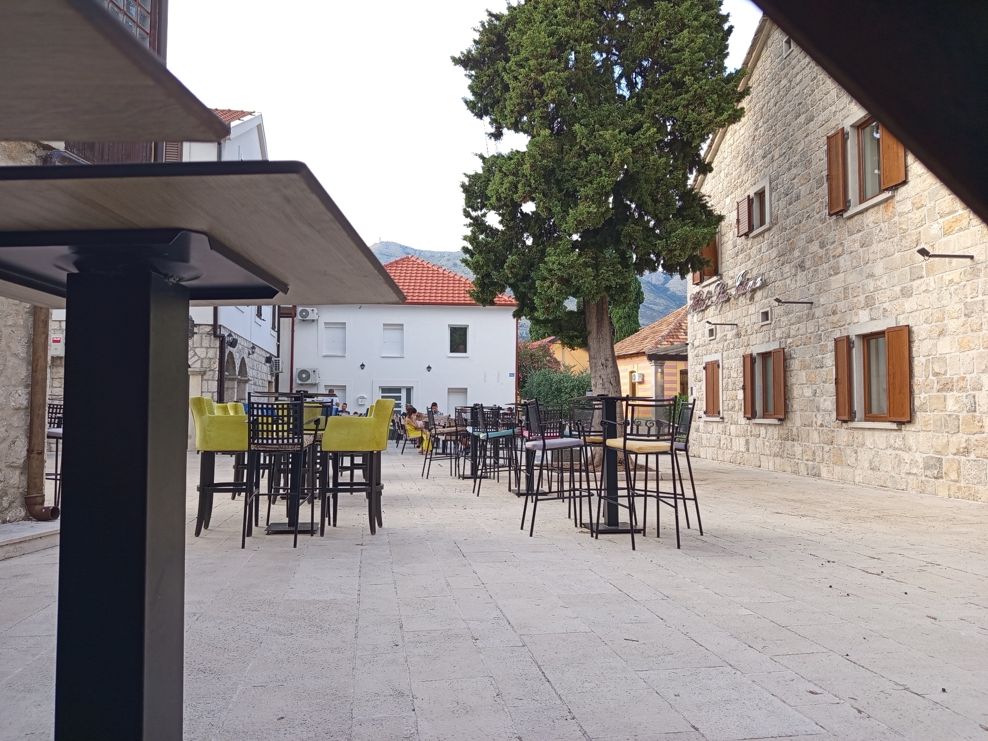 This is an outdoor shot of a seating area at what appears to be a cafe or restaurant in Trebinje, Bosnia and Herzegovina. The scene is set on a paved courtyard with a stone building on the right and a white building with red-tiled roofs in the background. A large, mature tree stands prominently in the middle of the scene, casting shadows.
The foreground is dominated by a dark, modern table and chairs, with the edge of the table and a pillar in focus. Beyond this, several high-top tables with dark metal frames and bright yellow bar stools are arranged, suggesting an outdoor dining or lounging area. Some of these tables have metal chairs with intricate designs. A few people are visible seated at some of the tables in the mid-ground, engaged in conversations or enjoying their time.
The architecture of the stone building features wooden shutters on its windows, and the white building has air conditioning units mounted on its exterior. The overall atmosphere suggests a pleasant, possibly late afternoon setting. The lighting is soft, indicating it's not midday. The weather appears to be fair, with a clear sky visible above the buildings and tree. The setting feels relaxed and inviting, typical of a European town square or a quiet cafe. No visible text is present in the image.