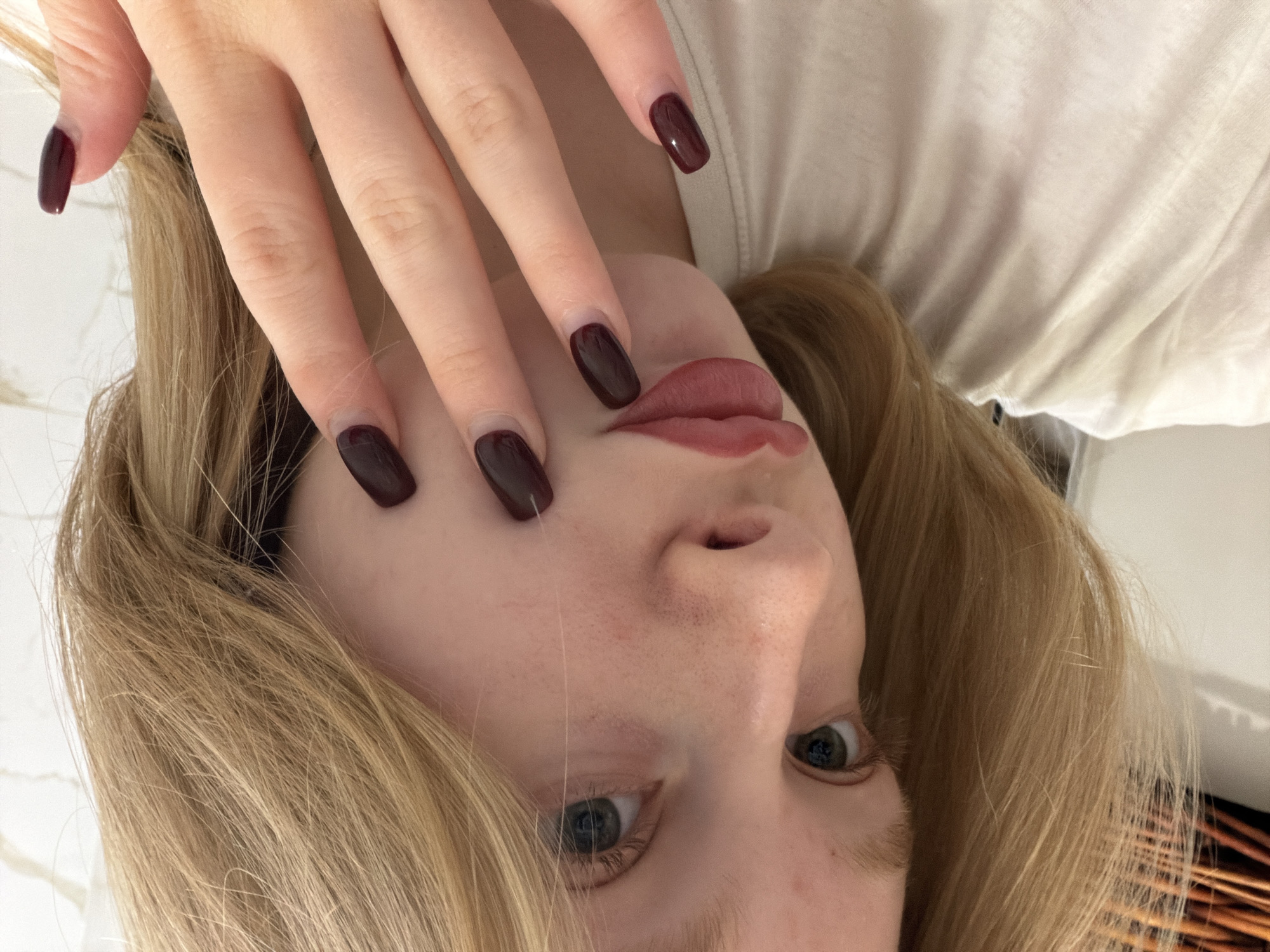 A close-up, upside-down selfie shows a fair-skinned woman with light brown hair framing her face. Her fingers, adorned with dark burgundy nail polish, partially obscure her face, especially around the left eye. Her other eye is visible and appears to be blue or grey. She wears lip color that is similar in color to her nail polish. The background is a white t-shirt and a white wall. There is no information to indicate the image's connection to the city of Rossiyskiy, Russia.
