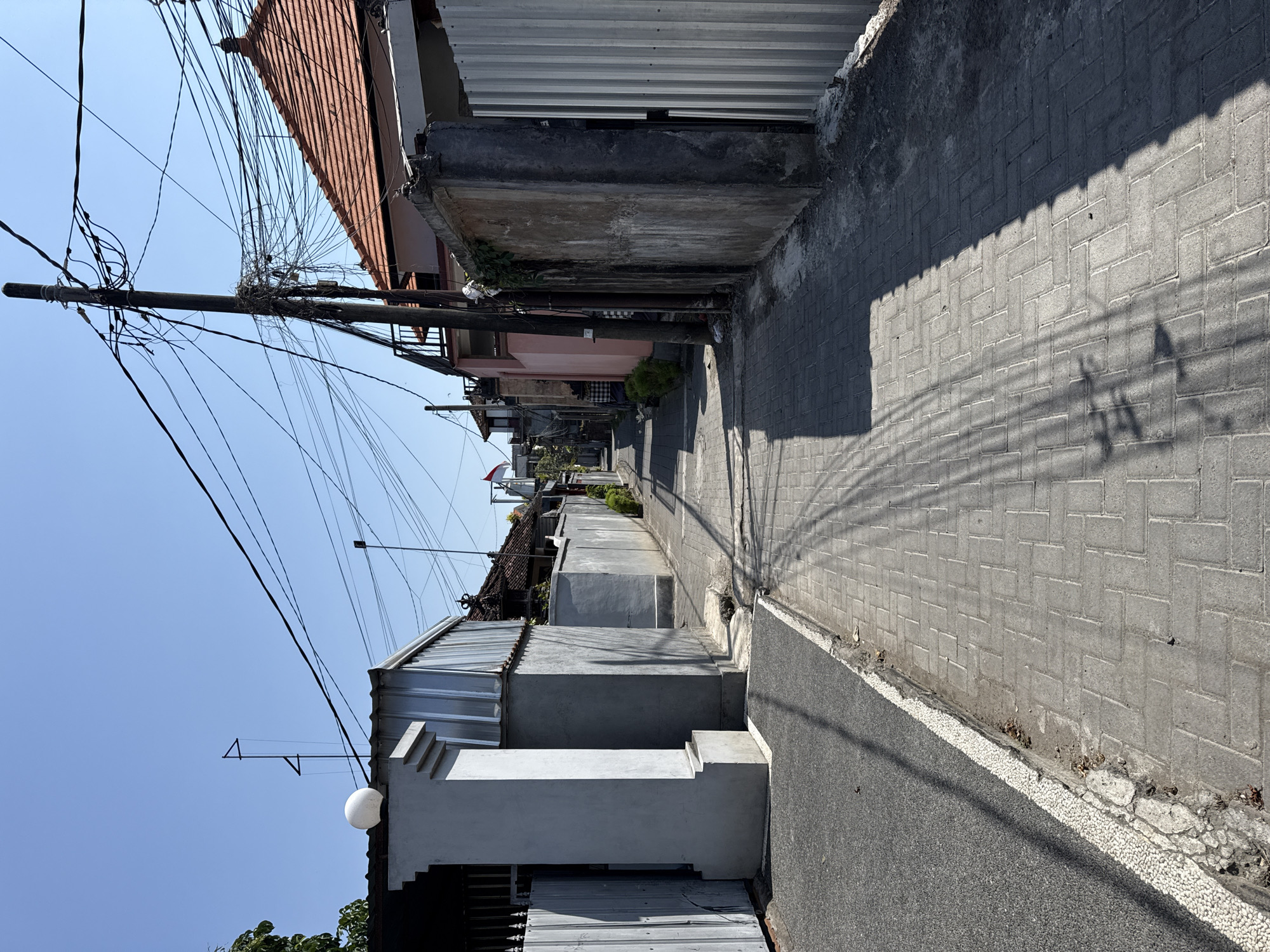 The image depicts a narrow, paved alleyway in Denpasar, Indonesia, likely in a residential or commercial district, on a clear, sunny day. The alley is lined with walls of varying materials, including concrete, corrugated metal, and plaster, some of which appear weathered. Buildings with tiled roofs are visible in the background. A prominent feature is a wooden utility pole heavily laden with a dense network of black electrical and telecommunication wires, casting intricate shadows on the brick-patterned pavement.
On the left side of the alley, a gray wall with a modern, white, spherical light fixture and a stepped architectural element is prominent. A dark gray, asphalt-like surface borders the paved walkway. Further down the alley, a red-tiled roof and a white and red Indonesian flag are visible. The right side of the alley features a rough, textured concrete wall and a section of a vertical, corrugated metal fence. Sparse green bushes are planted along some of the walls. The ground surface is primarily composed of gray interlocking pavers arranged in a herringbone pattern. The scene suggests a typical urban environment with overhead infrastructure and a mix of modern and older architectural styles. No people are visible in the image.