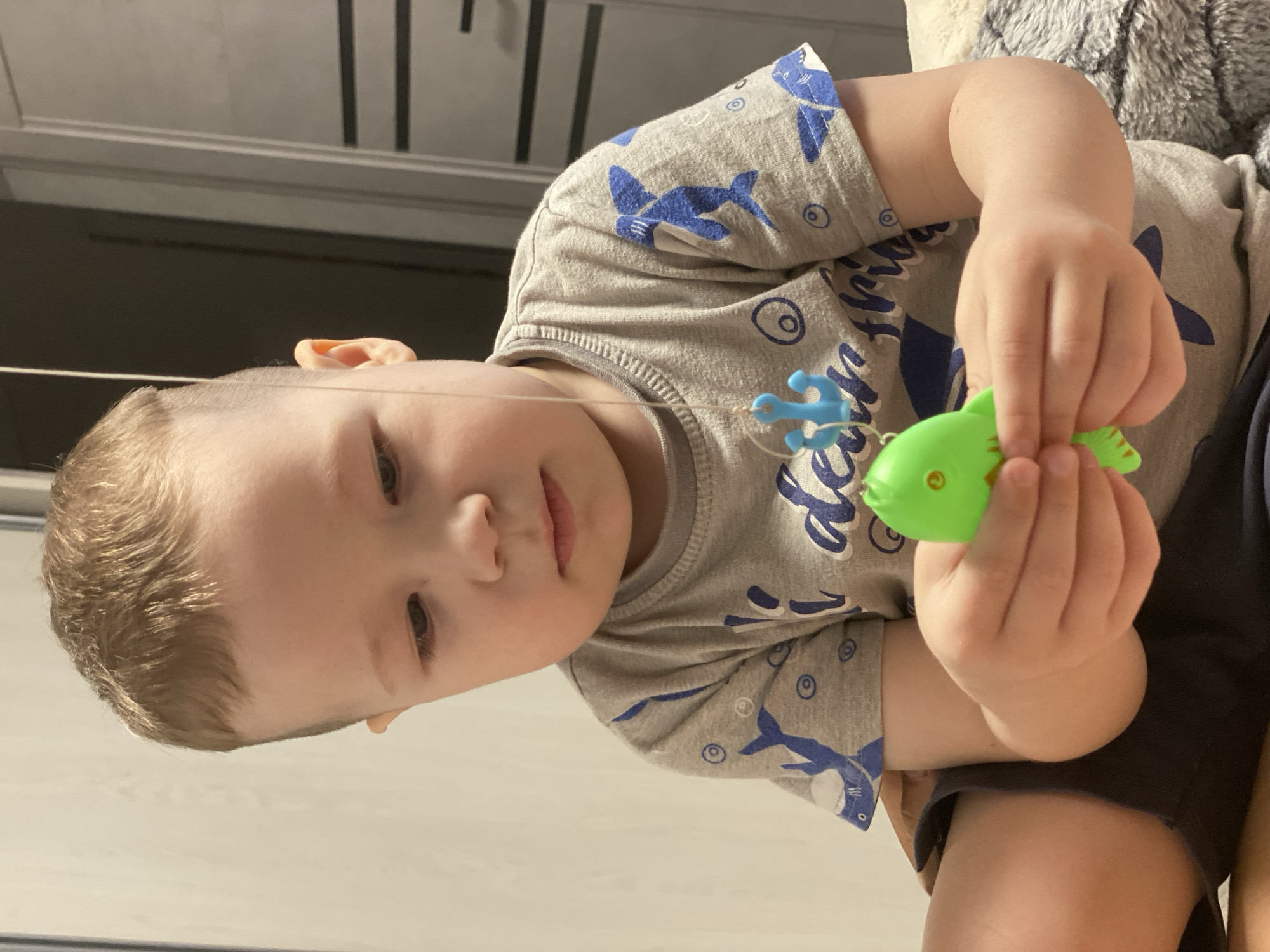A young child, a boy with light brown hair, is depicted indoors, engaged in playing with a toy. He is wearing a grey t-shirt adorned with blue shark and circular patterns, and dark shorts. The child is holding a bright green plastic toy fish in both hands, appearing focused downwards on the object. A thin white string extends from the toy fish, connecting to a smaller blue plastic object that resembles an anchor or starfish, suggesting it might be part of a magnetic fishing game.
The child's expression is neutral and concentrated on his play. The setting appears to be an indoor space, possibly a home in Sochi, Russia, given the provided context. The background is somewhat blurred but shows light-colored surfaces and a darker area, potentially furniture or another wall. A fuzzy, grey texture, possibly a blanket or plush toy, is visible near the child's upper right side. The lighting suggests a well-lit interior, either daytime or artificial illumination. There is no visible text in the image.