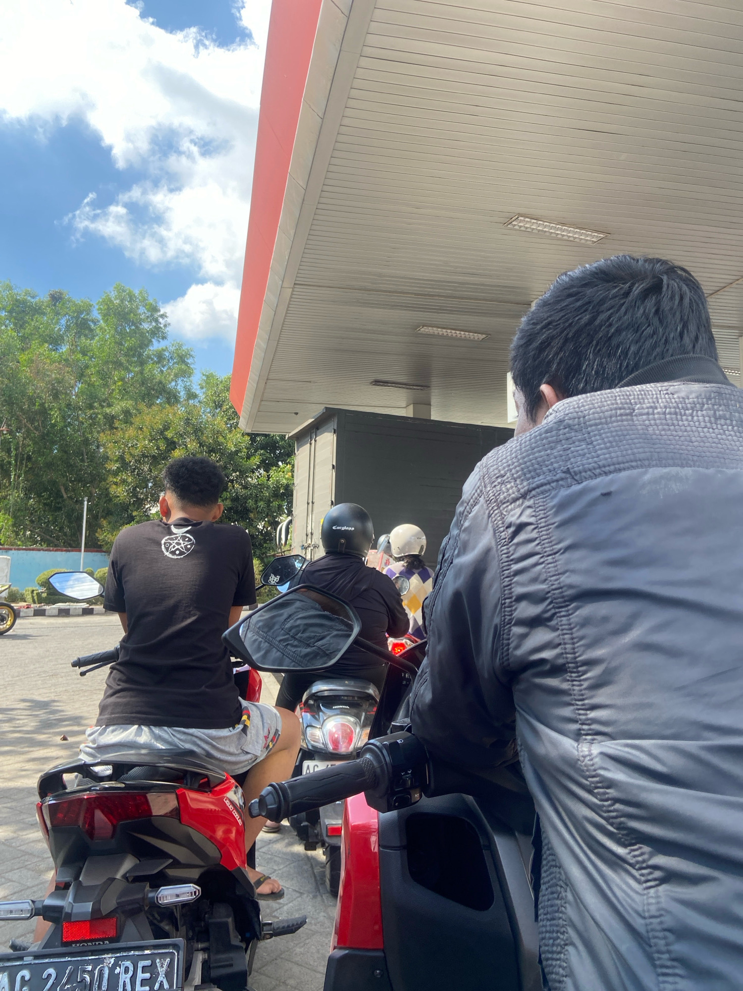 The image captures a scene at a gas station in Kedungwaru, Indonesia. In the foreground, two individuals are seated on motorcycles, facing away from the camera. The person on the left is wearing a black t-shirt with a graphic design and grey shorts. The person on the right is wearing a black puffer jacket. Behind them, other individuals on motorcycles are also visible, some wearing helmets. The gas station canopy, painted in white with a red trim, extends overhead. The background features lush green trees and a blue sky with scattered clouds, suggesting an outdoor, daytime setting. The overall atmosphere appears to be that of a typical busy gas station.