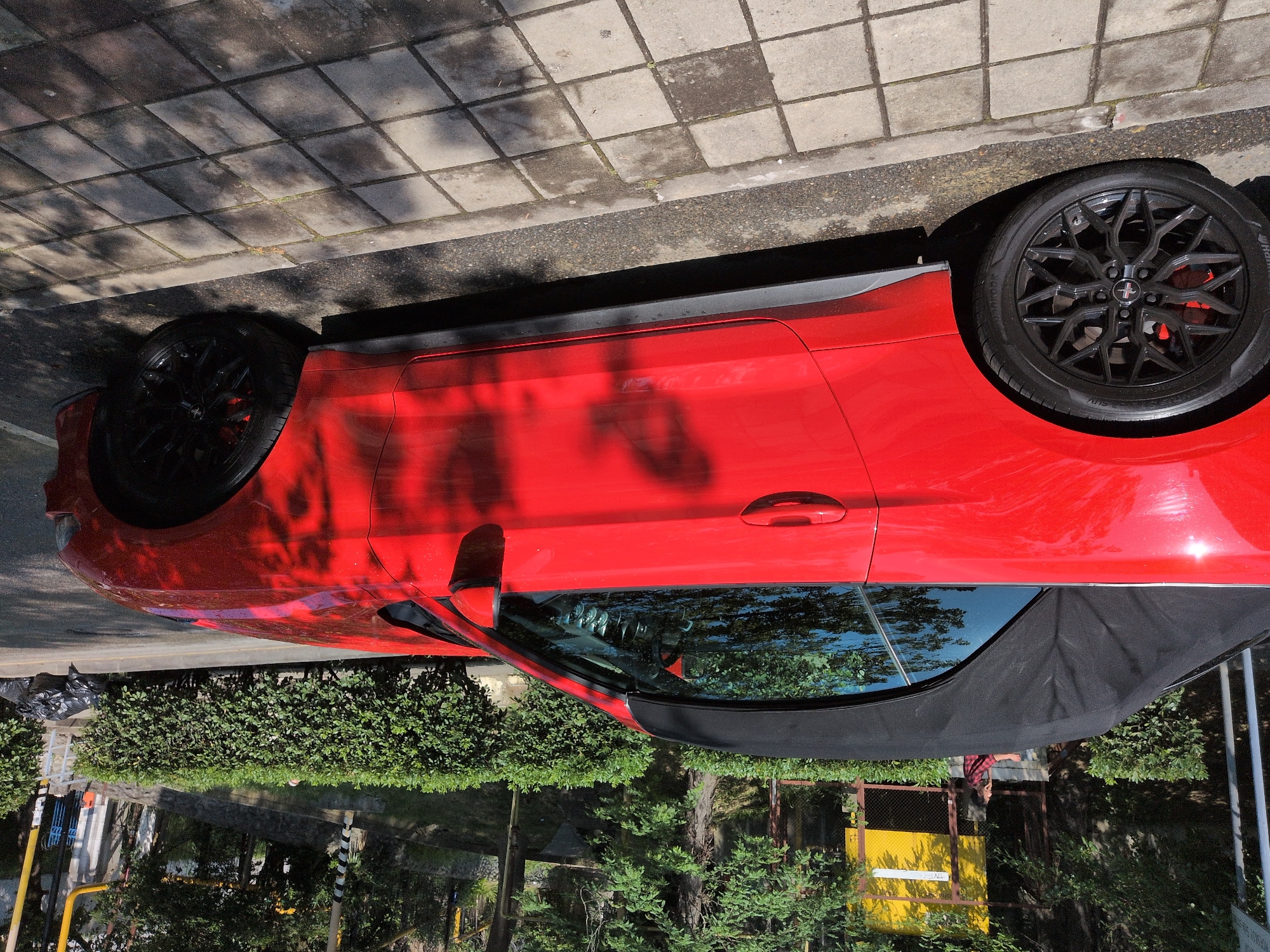 A bright red convertible is parked on a city street, likely in Sochi, Russia. The car, a Ford Mustang based on the emblem on the wheel, has its black soft top up. It's positioned parallel to a sidewalk paved with square concrete tiles, which are partially shaded by overhead foliage. Beyond the sidewalk, a manicured hedge runs along the side of the road, and behind that, trees and some urban infrastructure are visible. A person wearing a red and black plaid shirt is standing near the car, looking down at their phone. The scene appears to be a bright, sunny day, with shadows from trees dappling the car and the pavement.