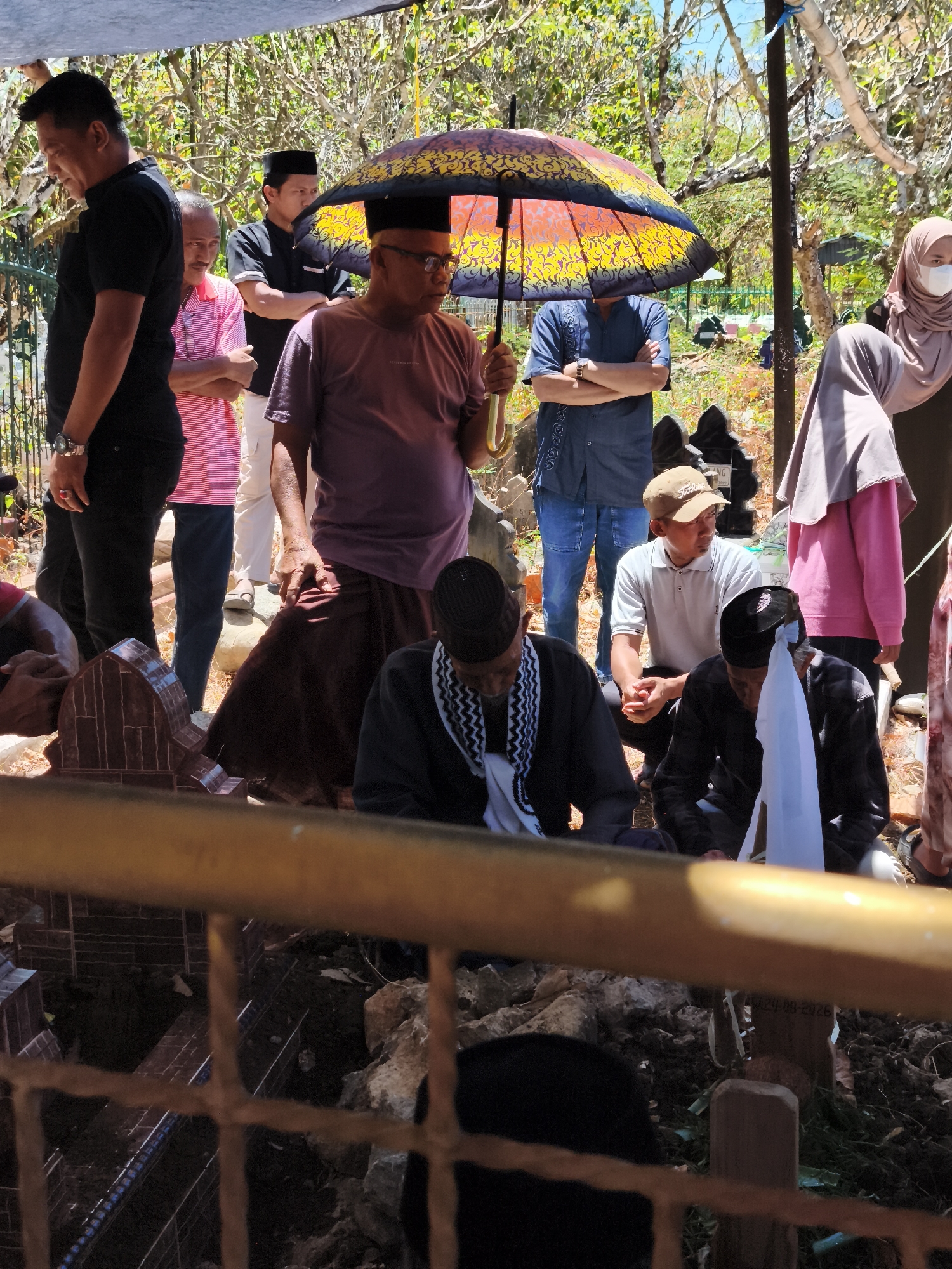 In Tonasa, Indonesia, a group of people are gathered at a gravesite under a makeshift canopy, shaded by a colorful umbrella. The scene appears somber, possibly a funeral or memorial service. Several individuals, primarily men, are dressed in dark clothing and some wear peci hats, common in Islamic cultural attire. One man, wearing glasses, stands prominently under the umbrella, holding it. Other men are seated or standing around the graves. Two women are visible in the background, one wearing a hijab and a face mask. The graves are marked with small, carved stone structures, and the ground is rough and uneven, with some rocks and foliage visible. The overall atmosphere is one of solemn reflection and community gathering at a place of remembrance.