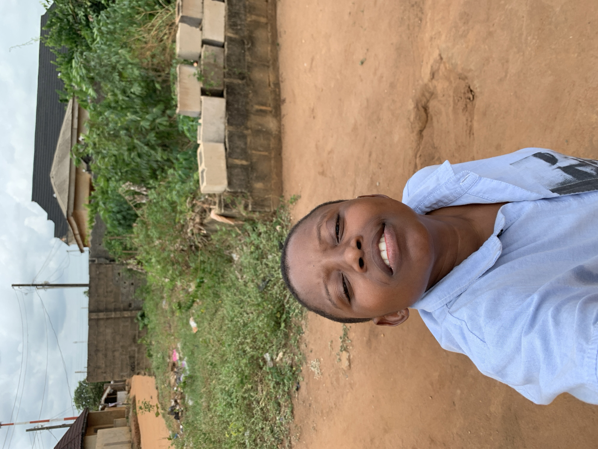 The image shows a person, likely a young adult, with dark skin and short hair, wearing a light blue collared shirt. They are smiling and appear to be taking a selfie. The background consists of a dirt path, vegetation including grass and trees, a concrete block wall, a building with a dark roof, and utility poles. The location is suggested to be Ikorodu, Nigeria, but there are no specific landmarks in the photo to confirm this. The individual is the primary subject of the image, and the surroundings suggest a residential or semi-urban environment.