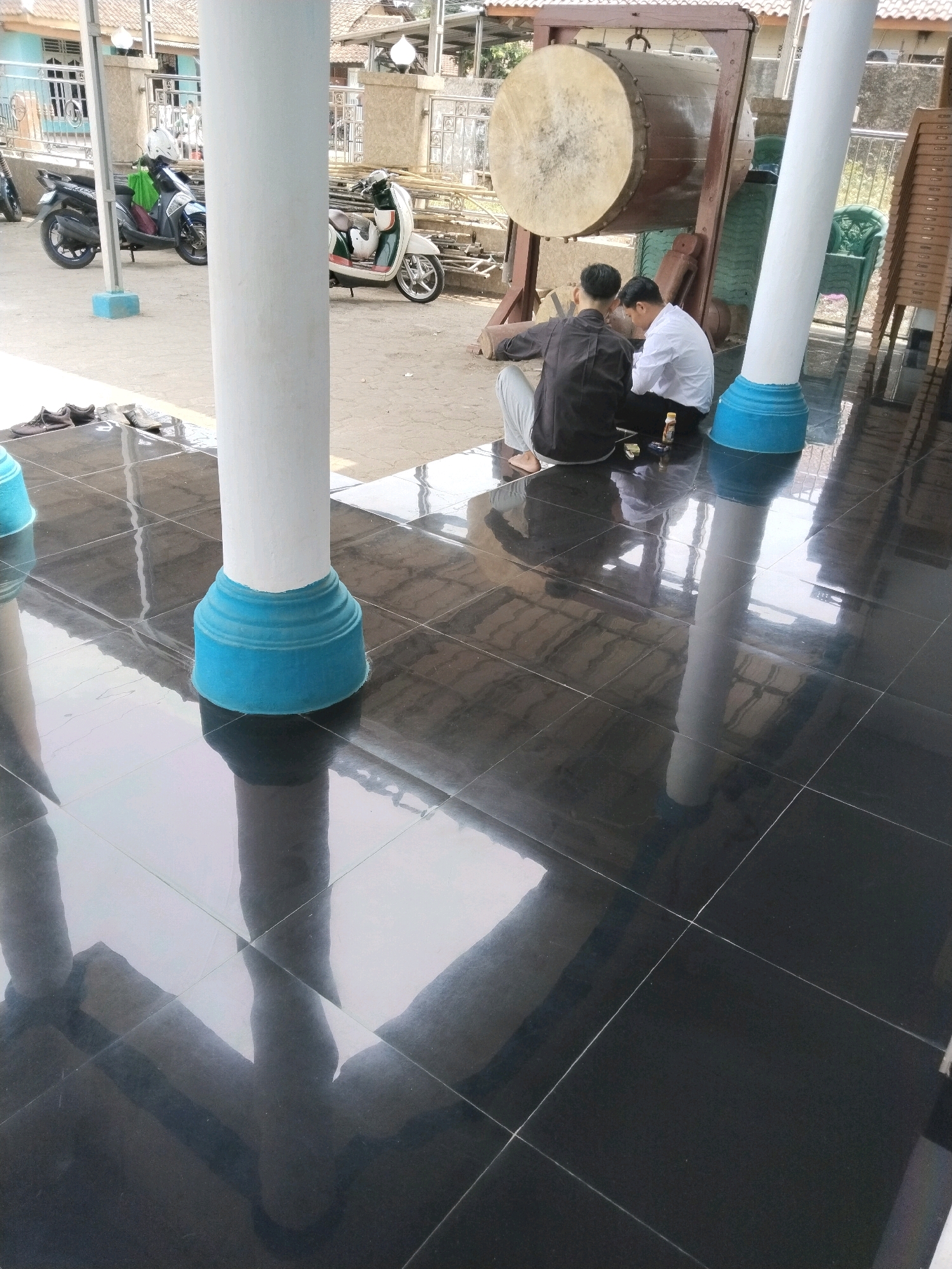 The image shows the exterior of a building, possibly a mosque or community hall, in Serang, Indonesia. In the foreground, two white pillars with blue bases stand on a reflective black tiled floor. Two young men, dressed in traditional Islamic attire, are seated on the floor in front of a large drum mounted on a wooden stand. The drum is round and appears to be made of wood and animal skin. The men are engaged in conversation or a quiet activity. In the background, there are several parked motorcycles, a green plastic chair, and stacks of brown chairs. The building itself appears to be made of light-colored materials, with windows and some decorative elements. The overall impression is a peaceful, communal outdoor space.