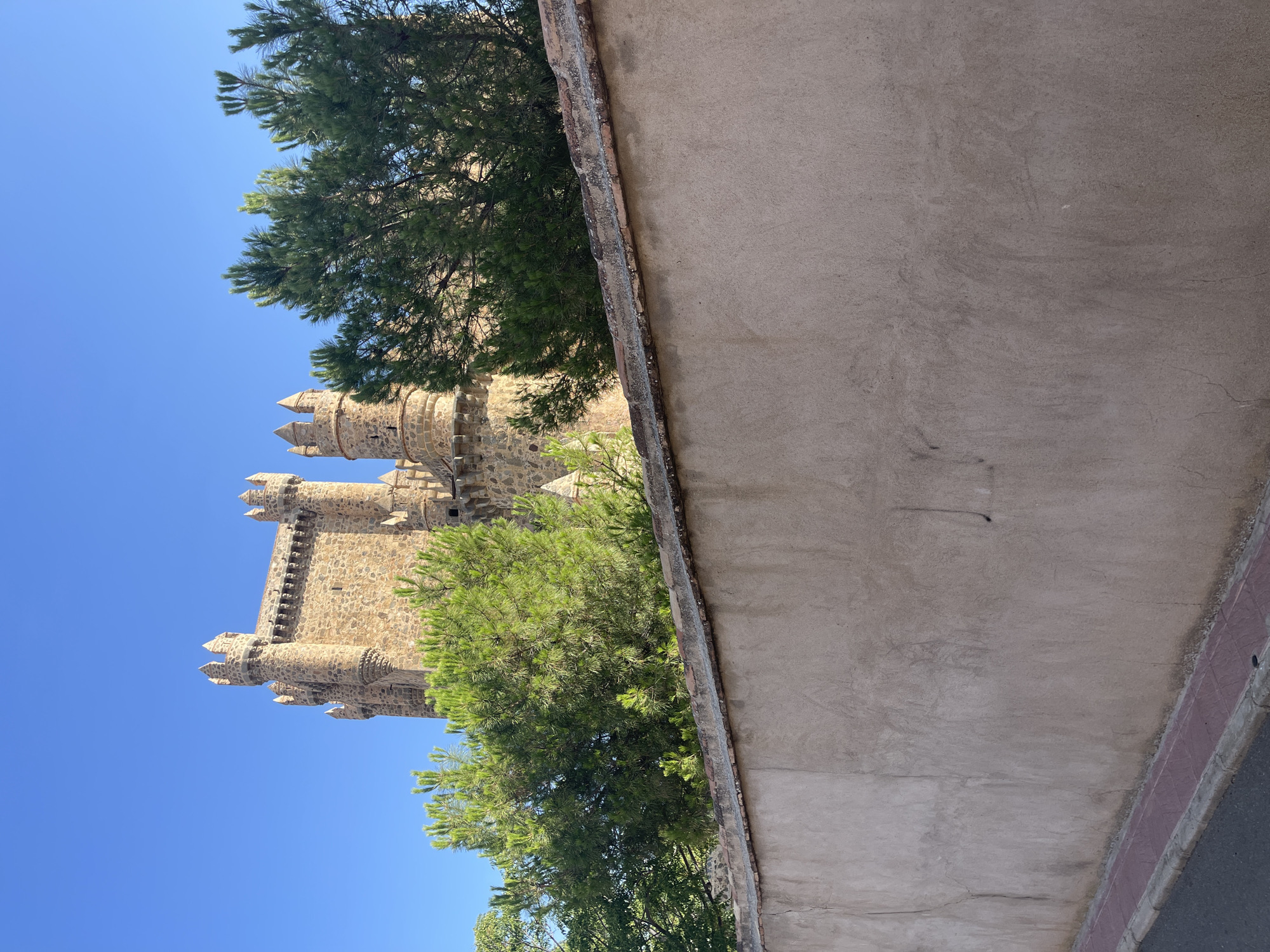 In Guadamur, Spain, a stone castle with crenellated towers rises against a clear blue sky, partially obscured by lush green pine trees. In the foreground, a weathered, beige stucco wall runs horizontally across the frame, topped with a band of weathered terracotta tiles. Below the wall, a narrow paved walkway with red tiles meets an asphalt road. The scene is static, with no people or discernible activity. The overall impression is one of historical architecture set within a natural environment under a bright, sunny day.