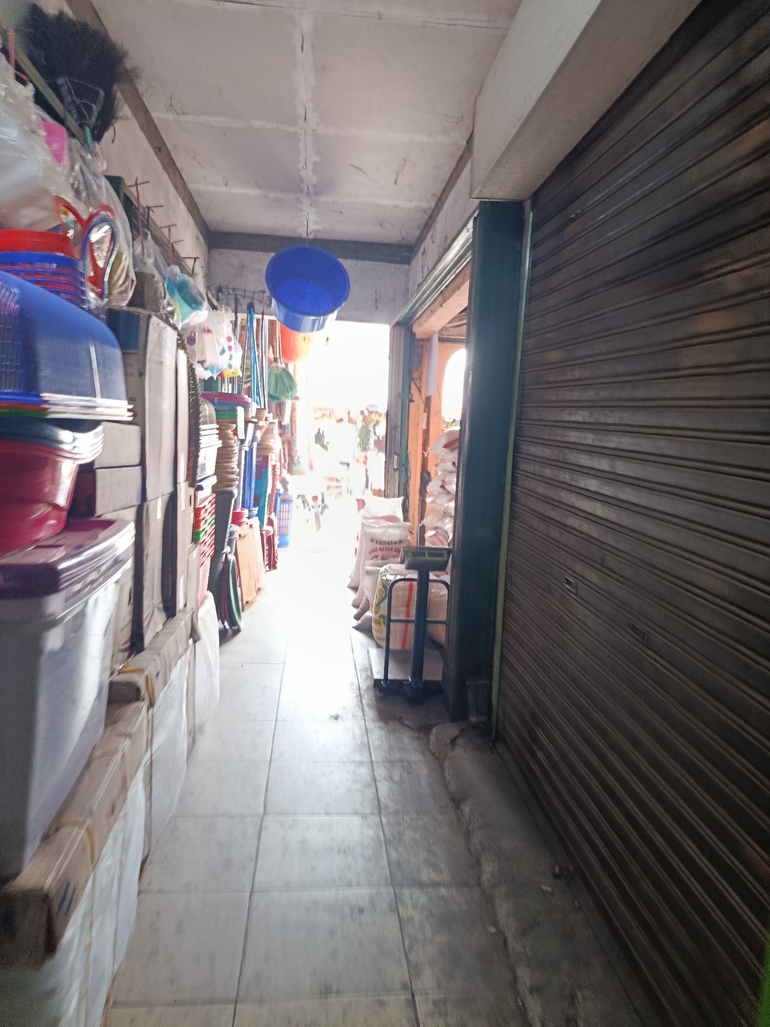 This is a photograph taken in Wanasari, Indonesia. The image depicts a narrow, covered market aisle lined with various goods. On the left, a stack of plastic containers, including blue and red tubs, is visible. Further down the aisle, more items are displayed, such as brooms, plastic baskets, and what appear to be cleaning supplies hanging from the ceiling. In the background, natural light streams in from an opening at the end of the aisle, illuminating sacks of goods and a weighing scale. On the right side of the aisle, a closed metal roll-up shutter dominates the view, suggesting a shop or stall. The floor is tiled. The overall impression is of a functional, busy marketplace, likely a retail area for household goods and other merchandise.
