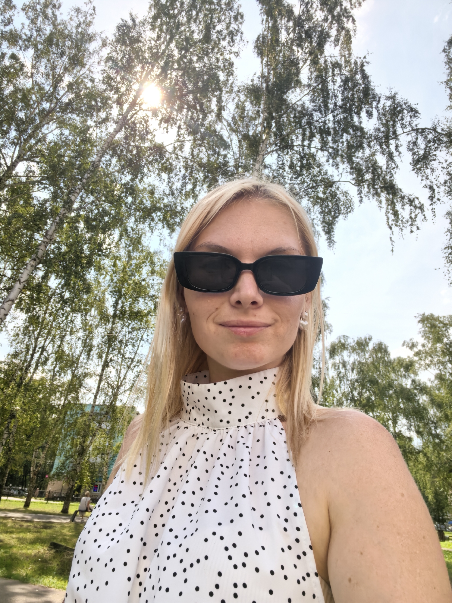 A medium shot, eye-level, outdoor selfie captures a blonde woman wearing black cat-eye sunglasses and a white halter top with black polka dots. She is positioned in the center of the frame, with her head and upper torso visible. The background is dominated by the lush green foliage of trees, with dappled sunlight filtering through the leaves and a bright sky overhead. In the distance, a light blue building and a park setting with a person sitting on a bench are visible. The image appears to be taken on a sunny day in the city of Sterlitamak, Russia.