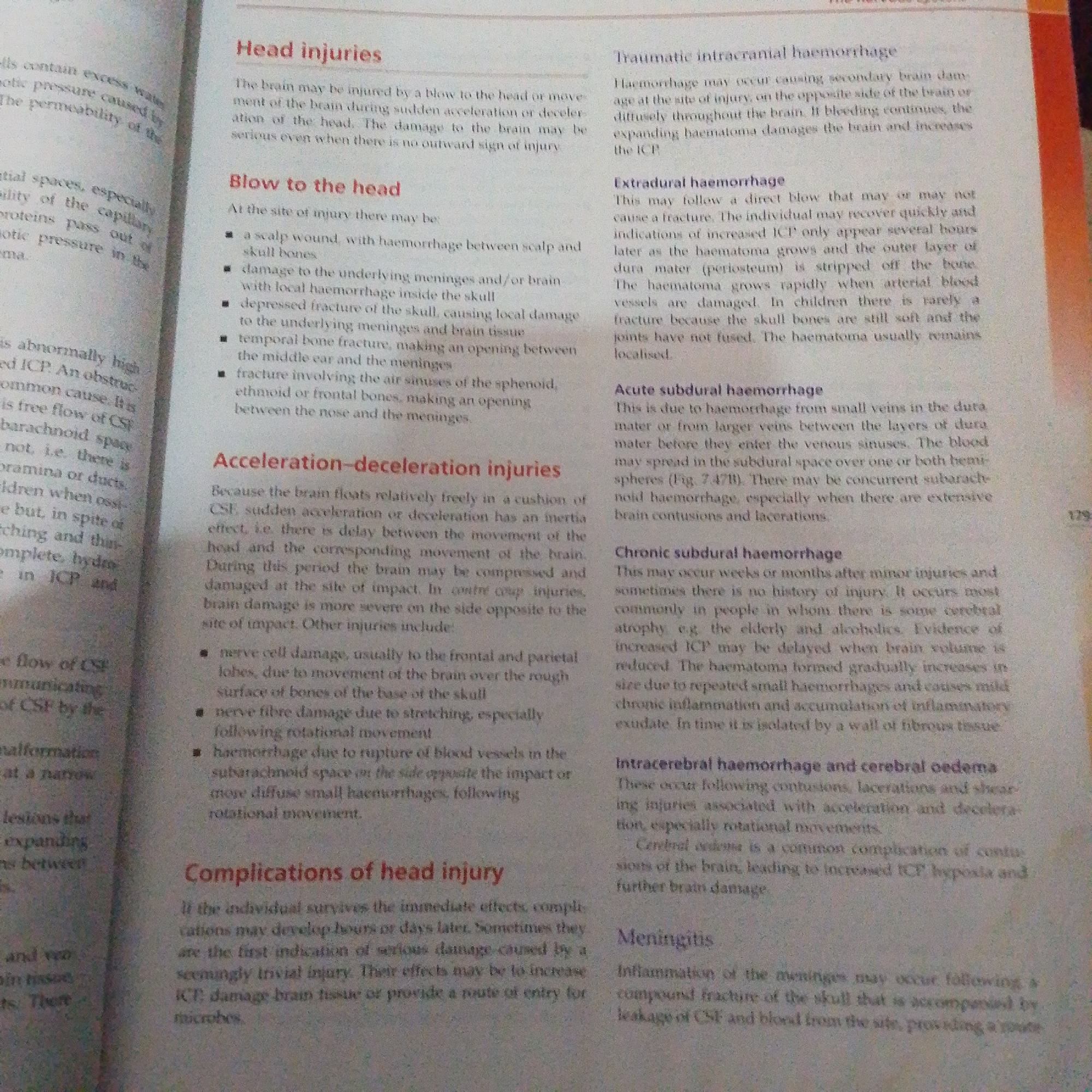 The image shows a close-up of an open textbook, viewed from a slightly angled perspective. The book is open to a page discussing "Head injuries" and related conditions like "Blow to the head," "Acceleration-deceleration injuries," "Complications of head injury," "Traumatic intracranial haemorrhage," "Extradural haemorrhage," "Acute subdural haemorrhage," "Chronic subdural haemorrhage," "Intracerebral haemorrhage and cerebral oedema," and "Meningitis." The text is printed in black on off-white paper, with headings in red. There is no indication of the time of day, weather, or specific emotions displayed. The setting appears to be indoors, likely a room where someone is studying or reading the book. No people are visible in the image. The text provides medical information related to head injuries.