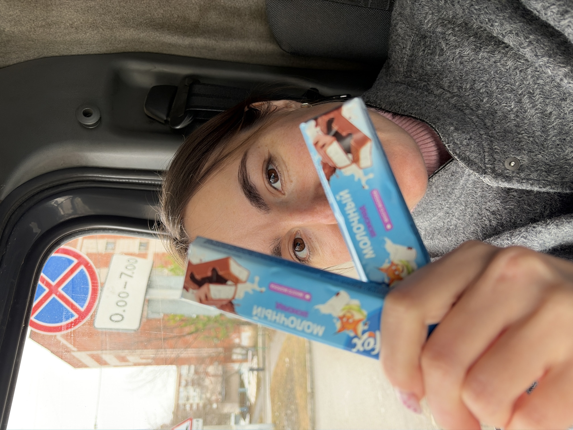 A person is inside a vehicle, holding two Fox "Molocnyy" (Milk) brand chocolate bars in front of their face. The person is wearing a grey coat and pink top. A parking prohibition sign is visible through the vehicle's window, along with a sign indicating parking restrictions from 0:00 to 7:00. The location is presumed to be in the city of Izhevsk, Russia, based on the Cyrillic script on the chocolate bar packaging.