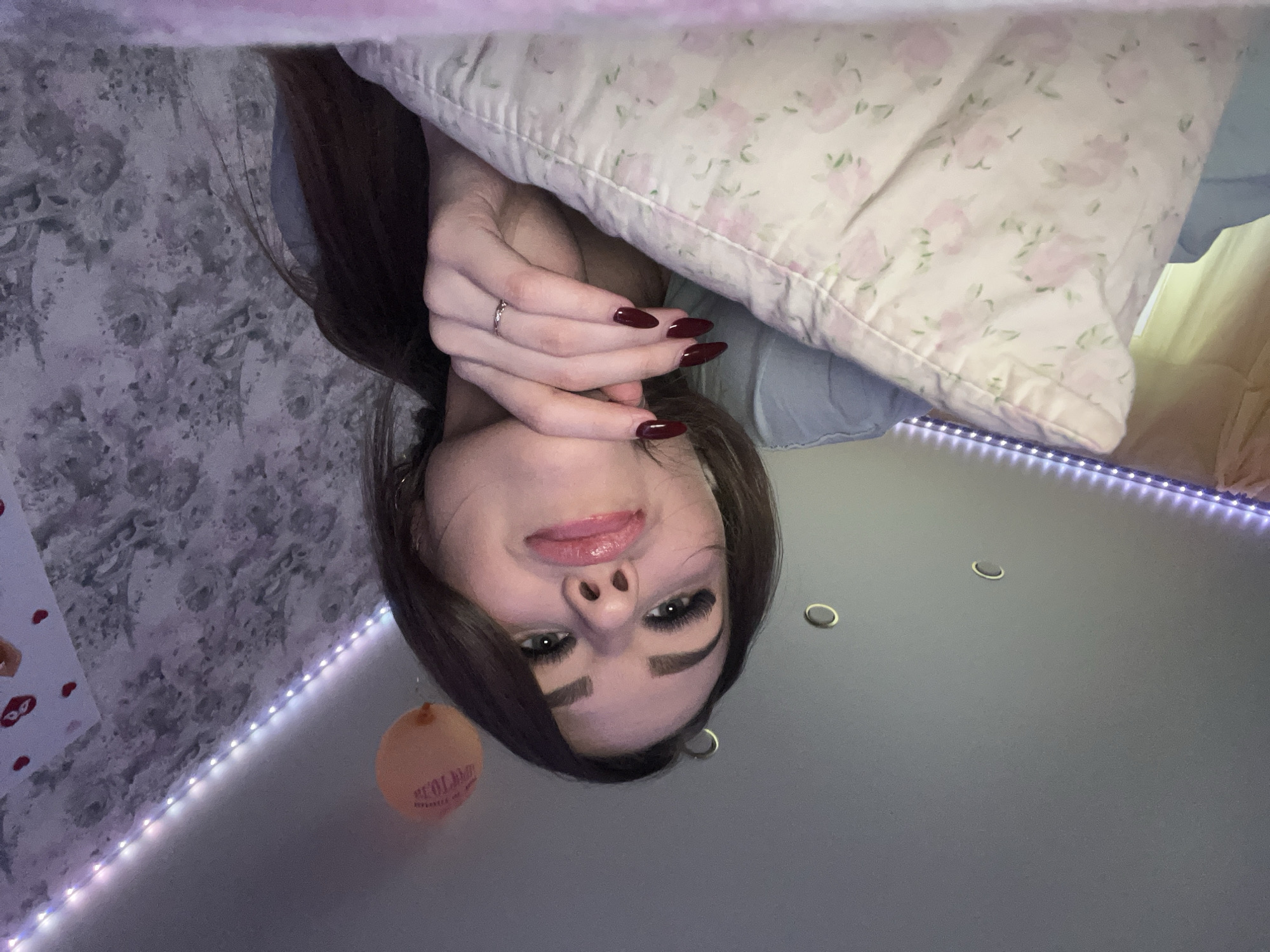 A young woman is upside down, looking at the camera. She has long brown hair and dark red nails. Her hands are near her face, and she is partially obscured by a pillow with a floral print. The background includes a gray wall with LED strip lights, a patterned wall, and several recessed lighting fixtures. A balloon is also visible. There is no evidence to suggest this image was taken in the city of Noviy, Russia.