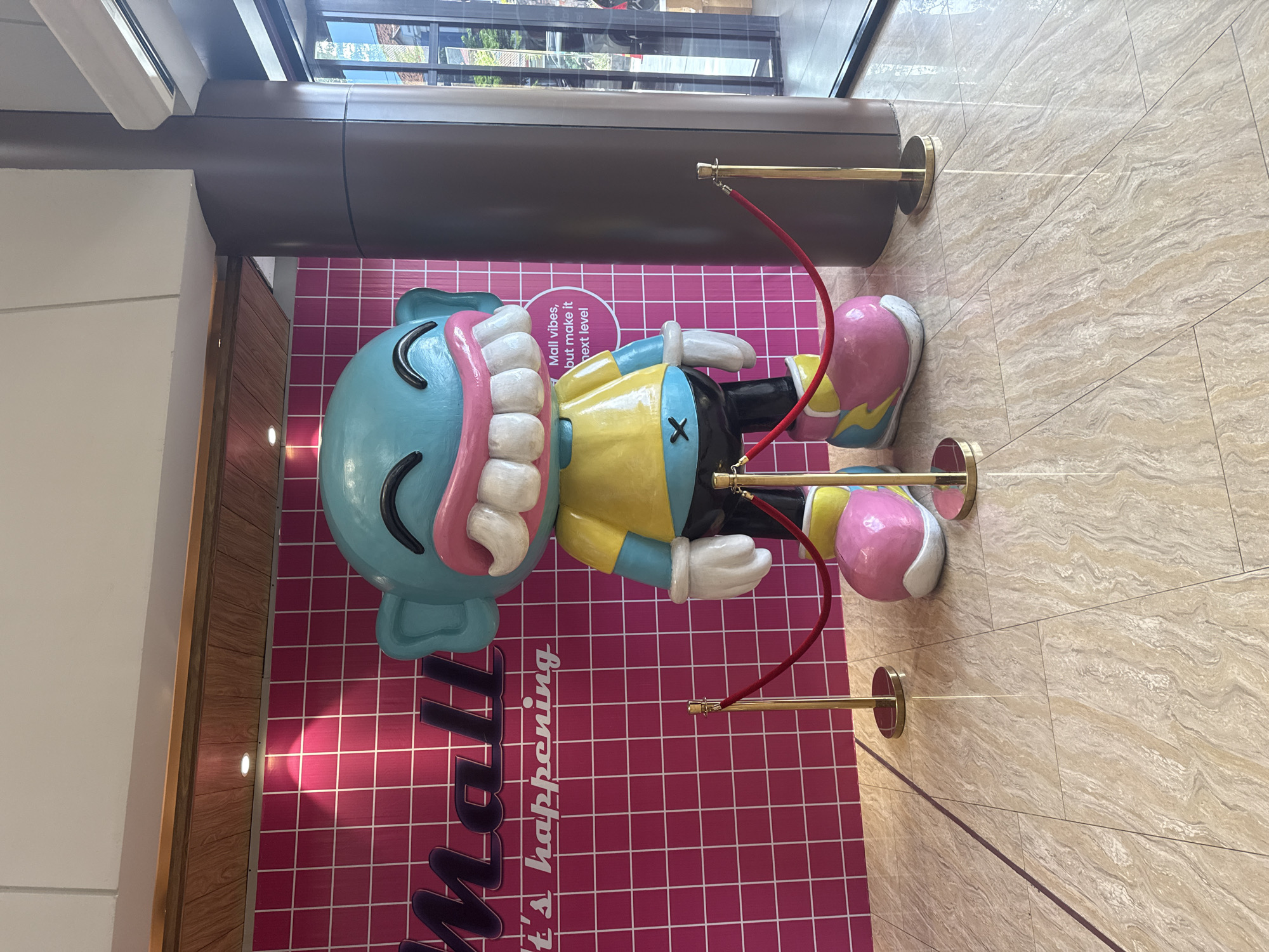 The image shows a brightly colored cartoon-style sculpture in what appears to be a shopping mall. The sculpture has a light blue head with a large, toothy grin, a yellow shirt with a black X on the chest, black pants, and mismatched pink and white shoes. The backdrop is a pink wall with a grid pattern and the words "Mall... It's happening." printed on it. The sculpture is cordoned off with a red rope attached to gold stanchions. The floor is light tan with a swirling pattern. The location is in Denpasar, Indonesia.