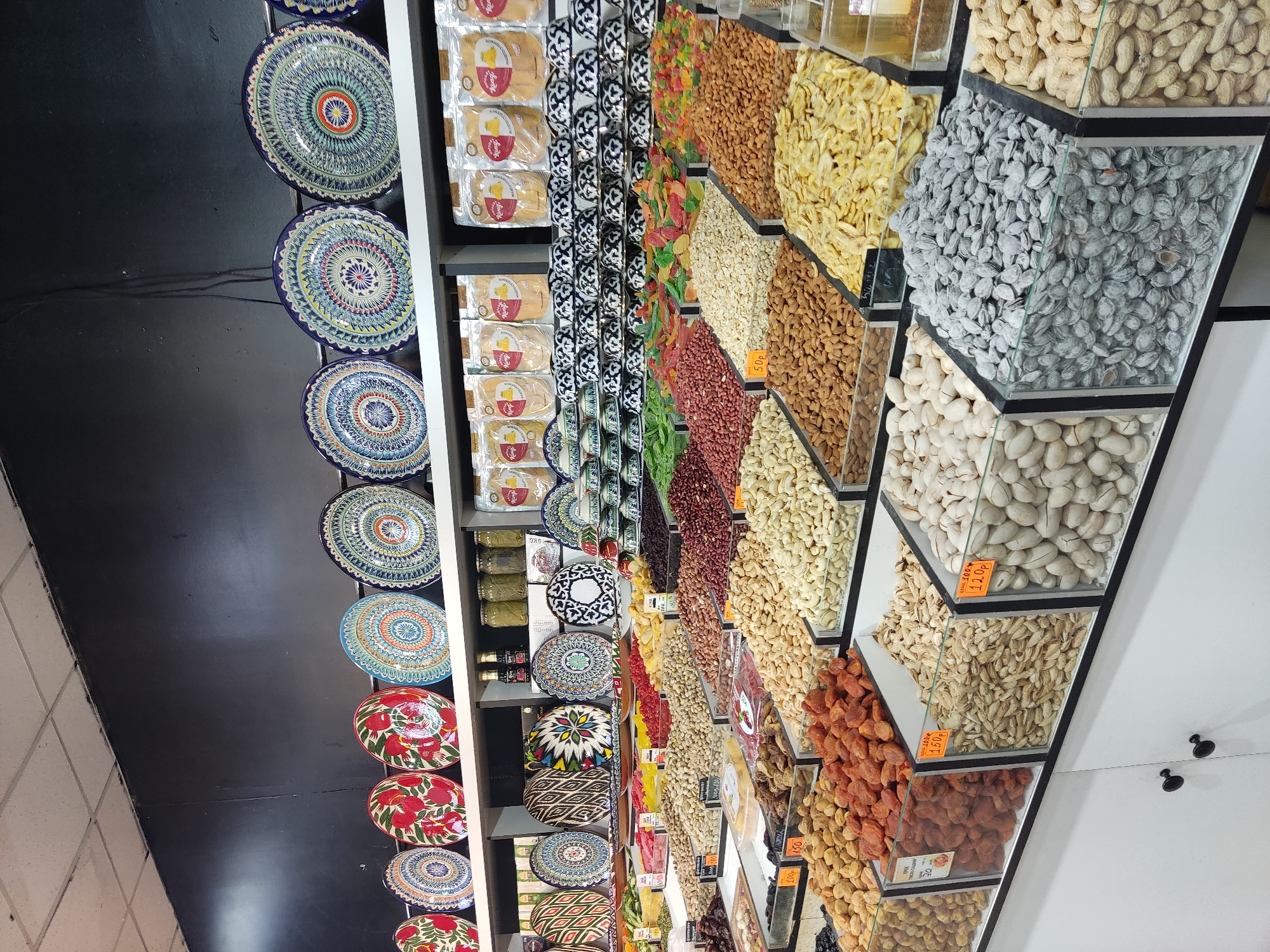 This image depicts a brightly lit interior of a market stall, likely a dried fruit and nut shop, showcasing a wide array of colorful products. In the foreground, large glass display bins are filled with various nuts and dried fruits. These include what appear to be pistachios, almonds, cashews, dried apricots, and possibly peanuts. Many of the bins have small price tags visible. Behind the nuts and fruits, there are shelves stacked with packaged goods, some of which appear to be snacks or food items in light-colored bags. Further back, decorative plates with intricate, colorful patterns are displayed on shelves, along with an assortment of small bowls or cups, also decorated. The overall environment suggests a retail setting focused on food products and decorative items. The image was taken in Noginsk, Russia.