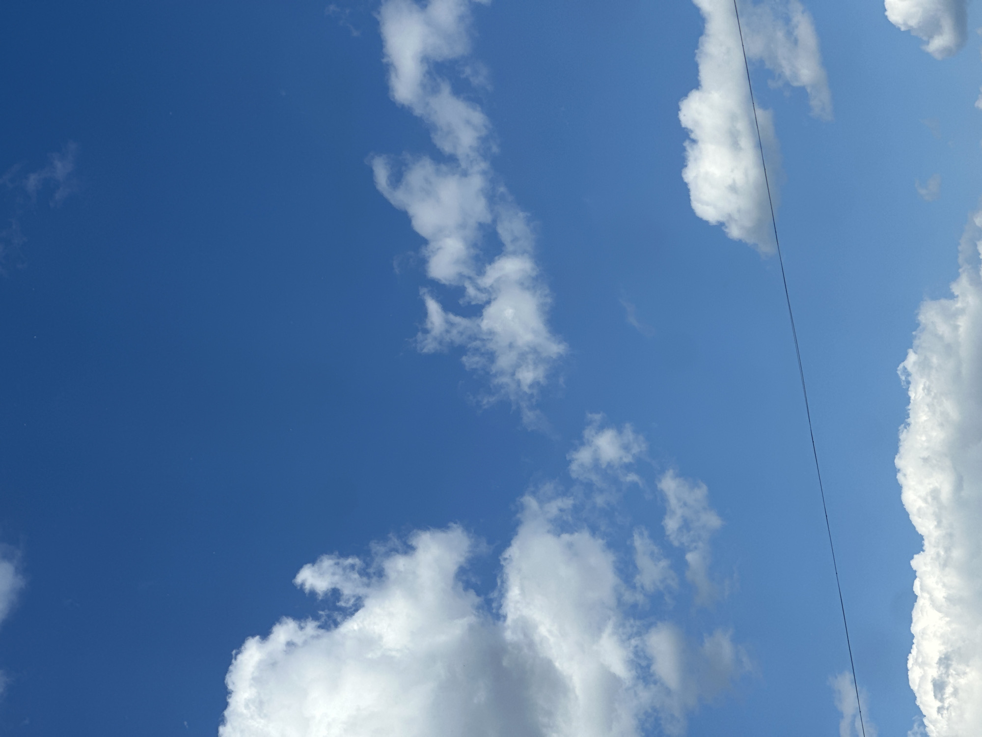 The image displays a clear blue sky with scattered white clouds. A thin, dark horizontal line, likely a power line, is visible across the lower half of the frame. No people or specific landmarks are discernible. The scene is consistent with an outdoor environment in Kazan, Russia, on a day with good visibility.