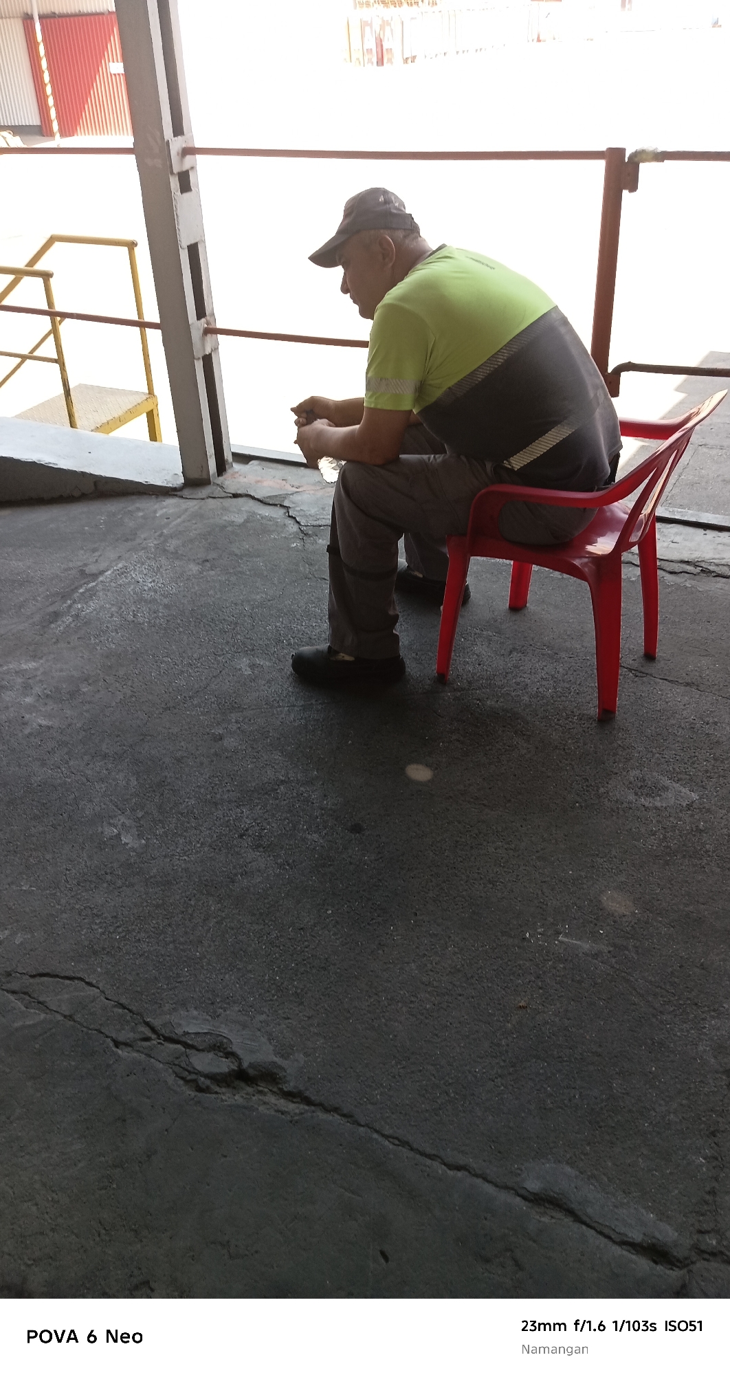 A man wearing a baseball cap and a green and grey t-shirt sits on a red plastic chair. He is looking down at his hands, which are clasped in his lap. He is wearing grey pants and black shoes. The man is sitting on a cracked asphalt surface. In the background, there is a metal railing and a building with red and white siding. The scene appears to be outdoors in an industrial or port area. The location is identified as Namangan City, Uzbekistan.