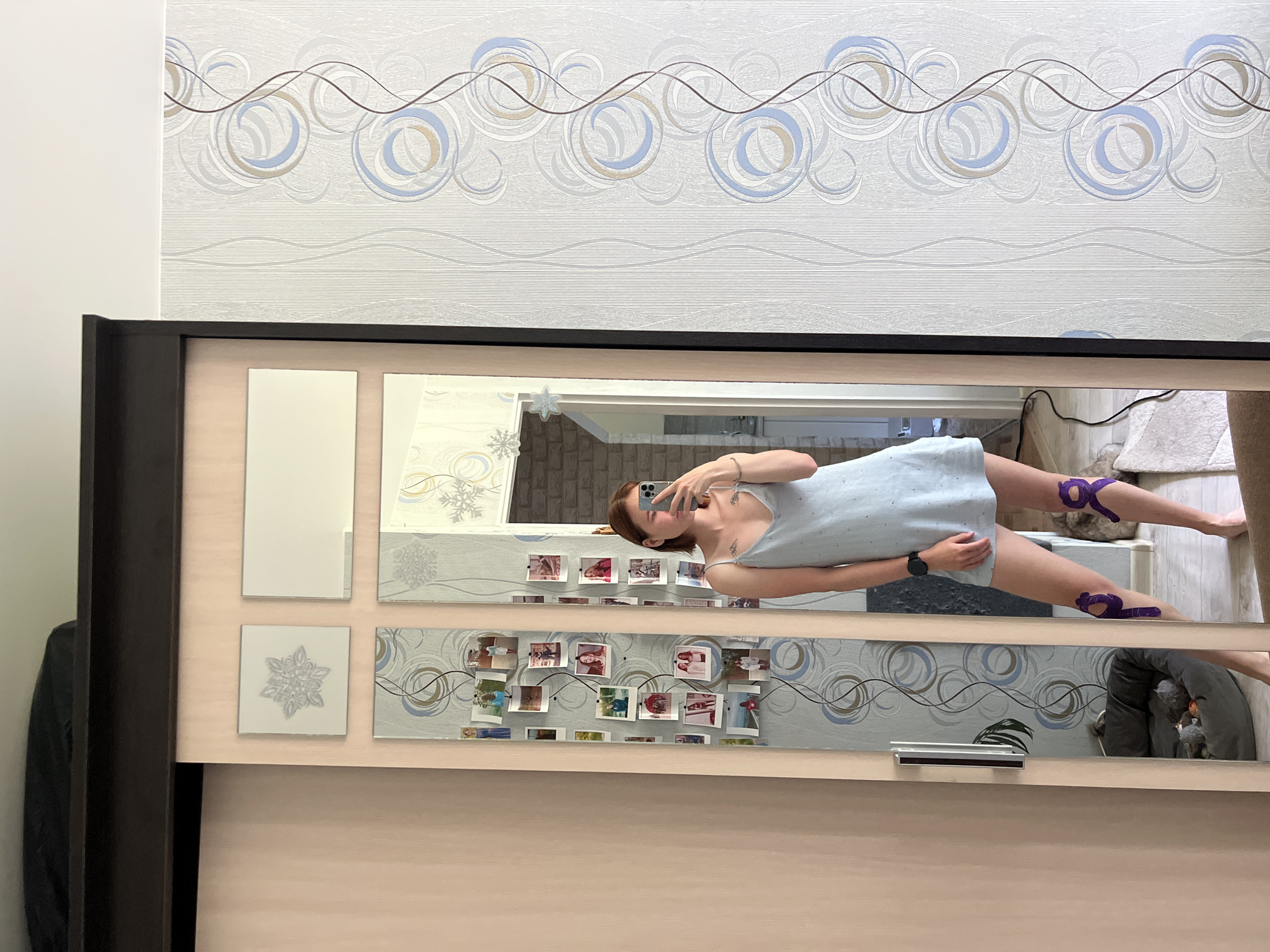 A young woman with short brown hair and fair skin is taking a selfie in a full-length mirror. She is wearing a light blue slip dress and has a watch on her left wrist. There are purple kinesiology tape applications on both of her knees. She is standing in what appears to be a bedroom or dressing area, with a large wardrobe with mirrored doors to her left. The wardrobe doors are decorated with framed pictures. The wall to the right of the wardrobe has a patterned wallpaper. In the reflection of the mirror, a dog bed with two dogs can be seen. The overall impression is that of a personal space, possibly in a residential apartment in Chaikovsky, Russia.