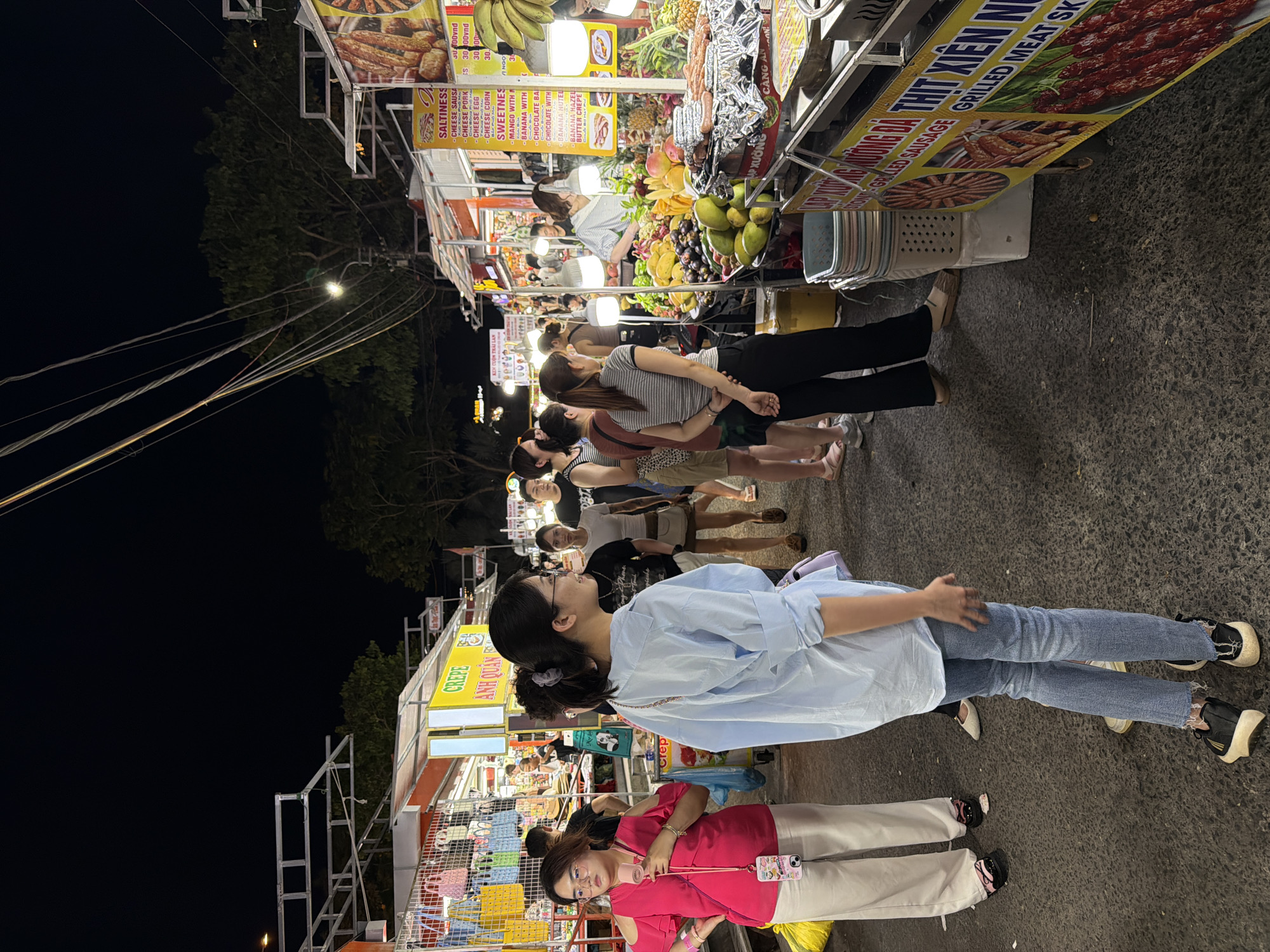 The image depicts a bustling night market scene in Đà Nẵng, Vietnam, likely on a street or pedestrian area. Several food stalls line both sides of a paved walkway, illuminated by overhead lights and the glow from the stalls themselves. People of various ages are seen browsing and interacting at the market.
On the left, a woman in a vibrant pink off-the-shoulder top and white pants holds a small pink fan, looking to her left. Beside her, another woman in a light blue button-down shirt and blue jeans stands facing away from the camera, her left hand slightly extended. Behind them, other shoppers and stall attendants are visible.
The stalls offer a variety of items, including what appears to be handbags and accessories on the left, and a prominent fruit stall on the right filled with an assortment of colorful fruits like mangoes, grapes, and pineapples. Next to the fruit stall is a grill with sausages cooking. Signs in Vietnamese are visible on the stalls, advertising food items such as "CREPE," "SALTINESS," "SWEETNESS," "GRILLED SAUSAGE," and "GRILLED MEAT SKEWER."
The overall atmosphere is lively and dynamic, characteristic of a popular evening market. The ground is asphalt, and overhead, power lines and streetlights are visible against a dark night sky. The composition suggests a candid snapshot capturing the energy of the location.