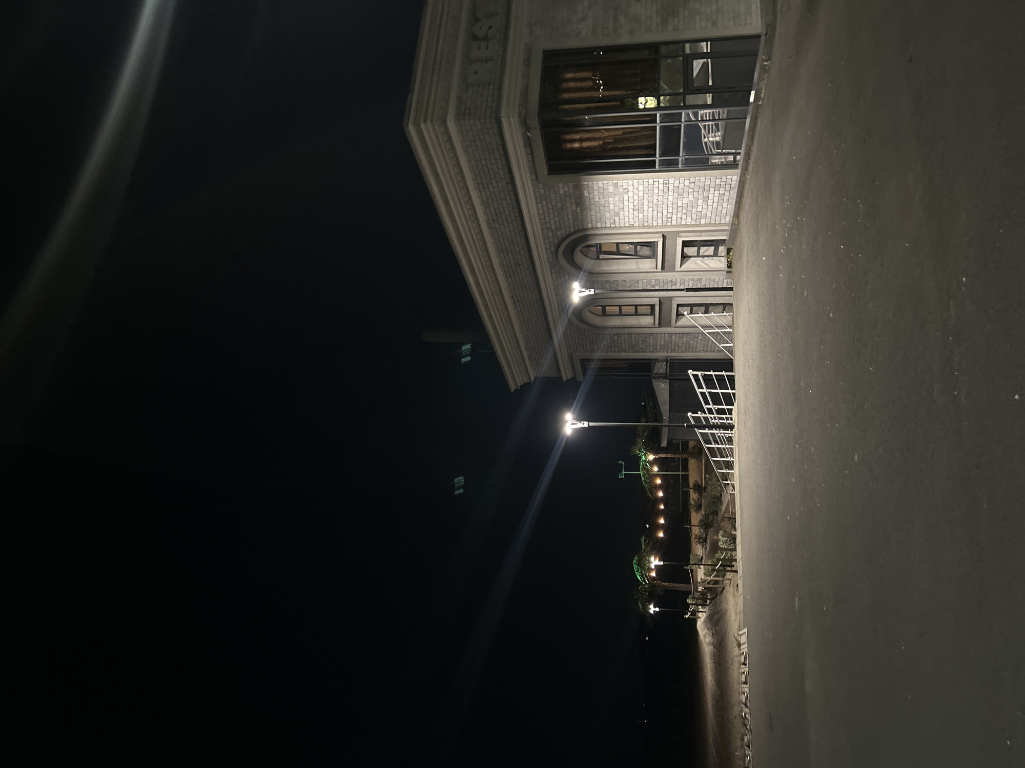 The image depicts a nighttime scene in Burgas, Bulgaria. The frame is dominated by a grey building with a brick facade on the right side, with the word "REST" visible at the top of the building. The building has large windows and arches. Two lampposts illuminate the front of the building, creating bright spots in the dark scene. A metal railing runs along the side of the building. Further down, there are palm trees decorated with lights along the coast. The left side of the image is primarily a dark sky, with a blurred light source at the top. The street surface is visible on the right, and the beach is visible at the bottom.