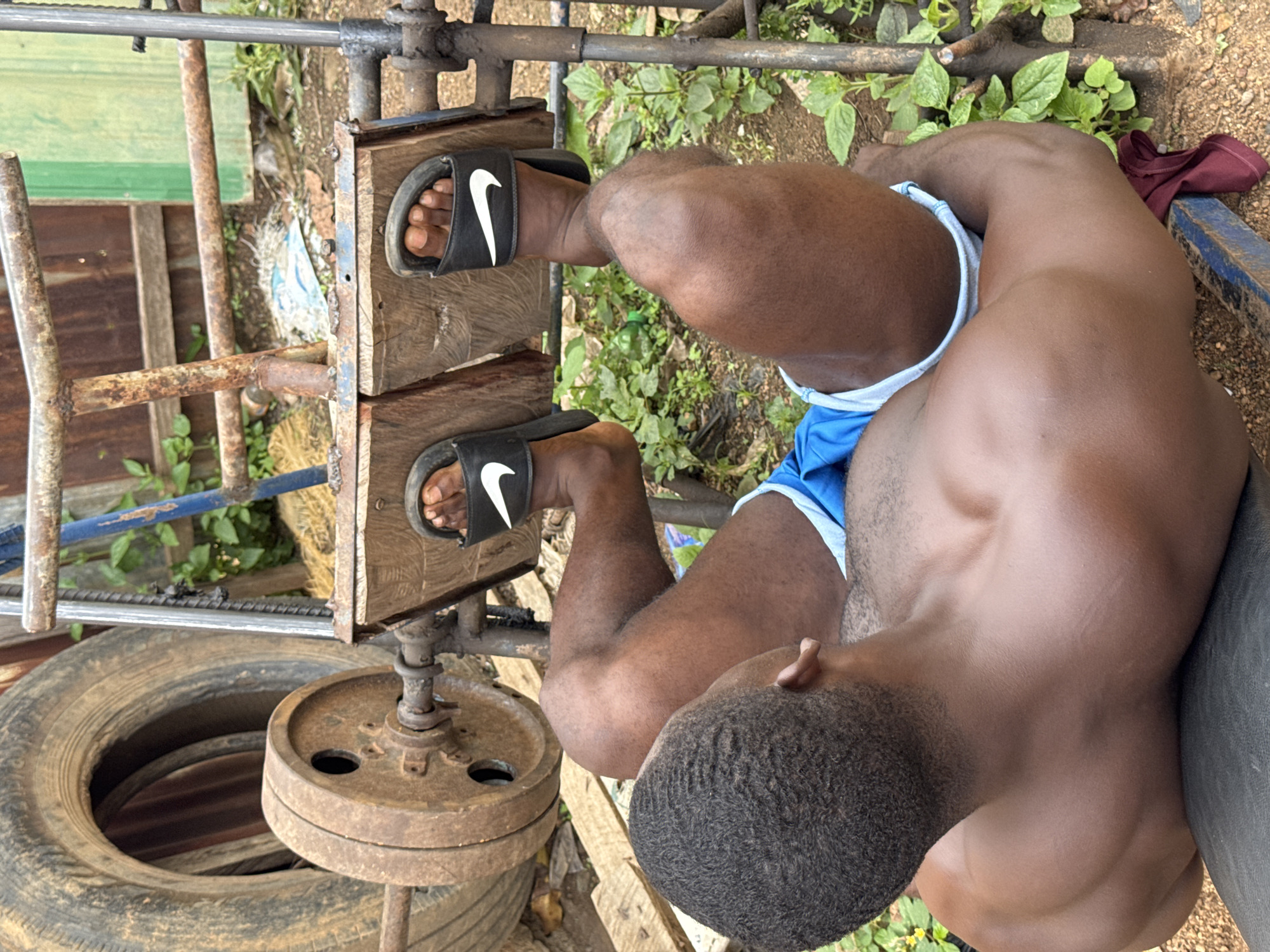 This media file shows a Black man exercising with a makeshift weightlifting machine in an outdoor setting. The machine appears to be constructed from scrap metal, wood, and car tires, with rusty metal rods and plates serving as weights. The man is wearing black Nike slides and blue shorts, with his bare chest visible. He is seated on a wooden bench, with his feet resting on the machine. The scene is set outdoors, with greenery and dirt visible in the background, suggesting a rural or semi-rural location in Akwanga, Nigeria. The lighting suggests it is daytime, possibly late afternoon due to the warm tones. There are no visible expressions of emotion, but the focus on the exercise equipment and the man's physique implies a dedication to physical training. No text is visible in the image.