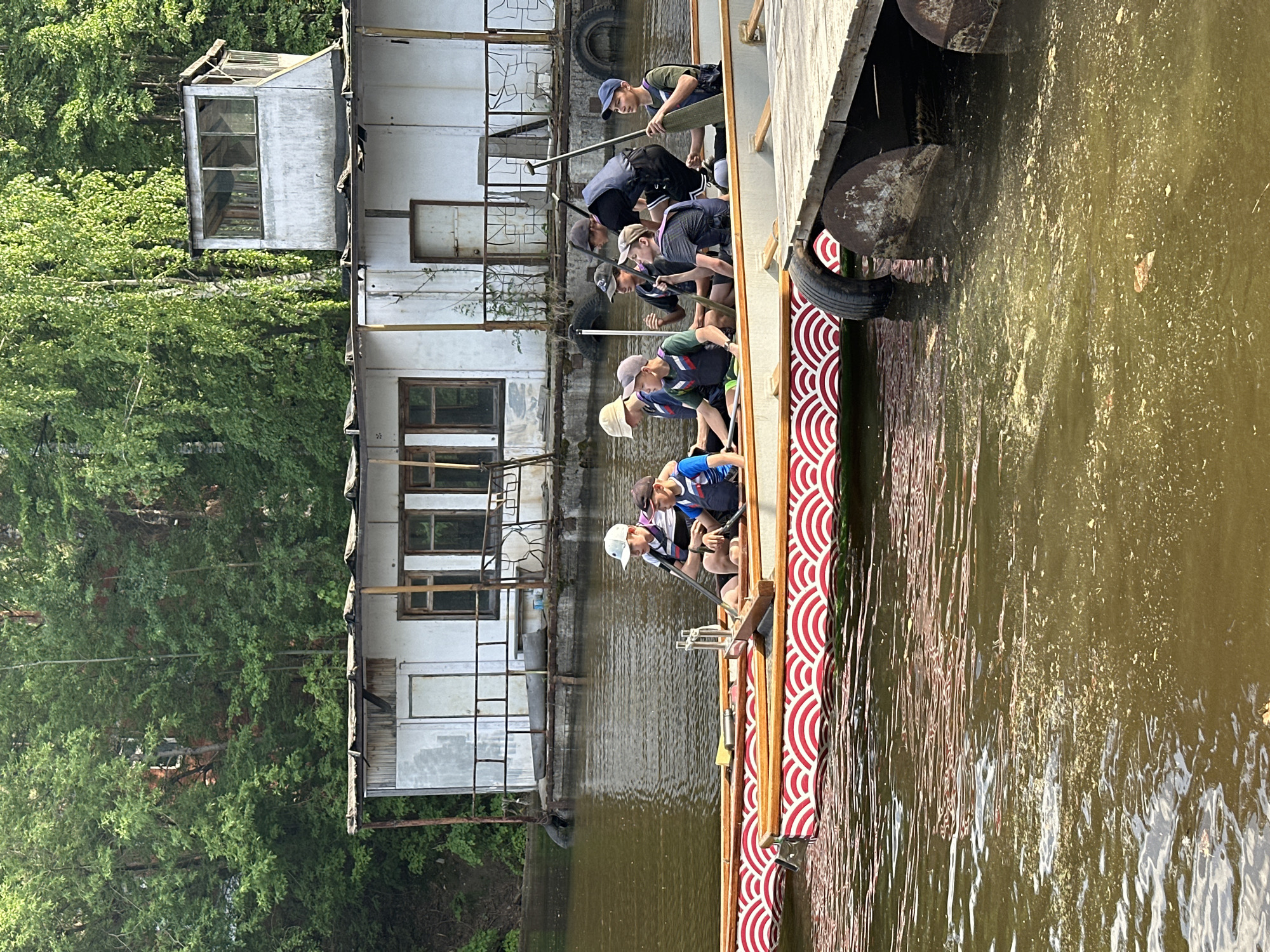 A group of about eight young people are rowing on a river in Chaikovsky, Russia. They are in a long, narrow boat with a red and white patterned side. The river is murky and reflects the surrounding trees and buildings. The people are all wearing different types of clothing, but most of them have caps on. A dilapidated building with multiple doors and windows stands next to the riverbank, partially obscured by dense trees. Old tires hang from the building and the edge of the boat.