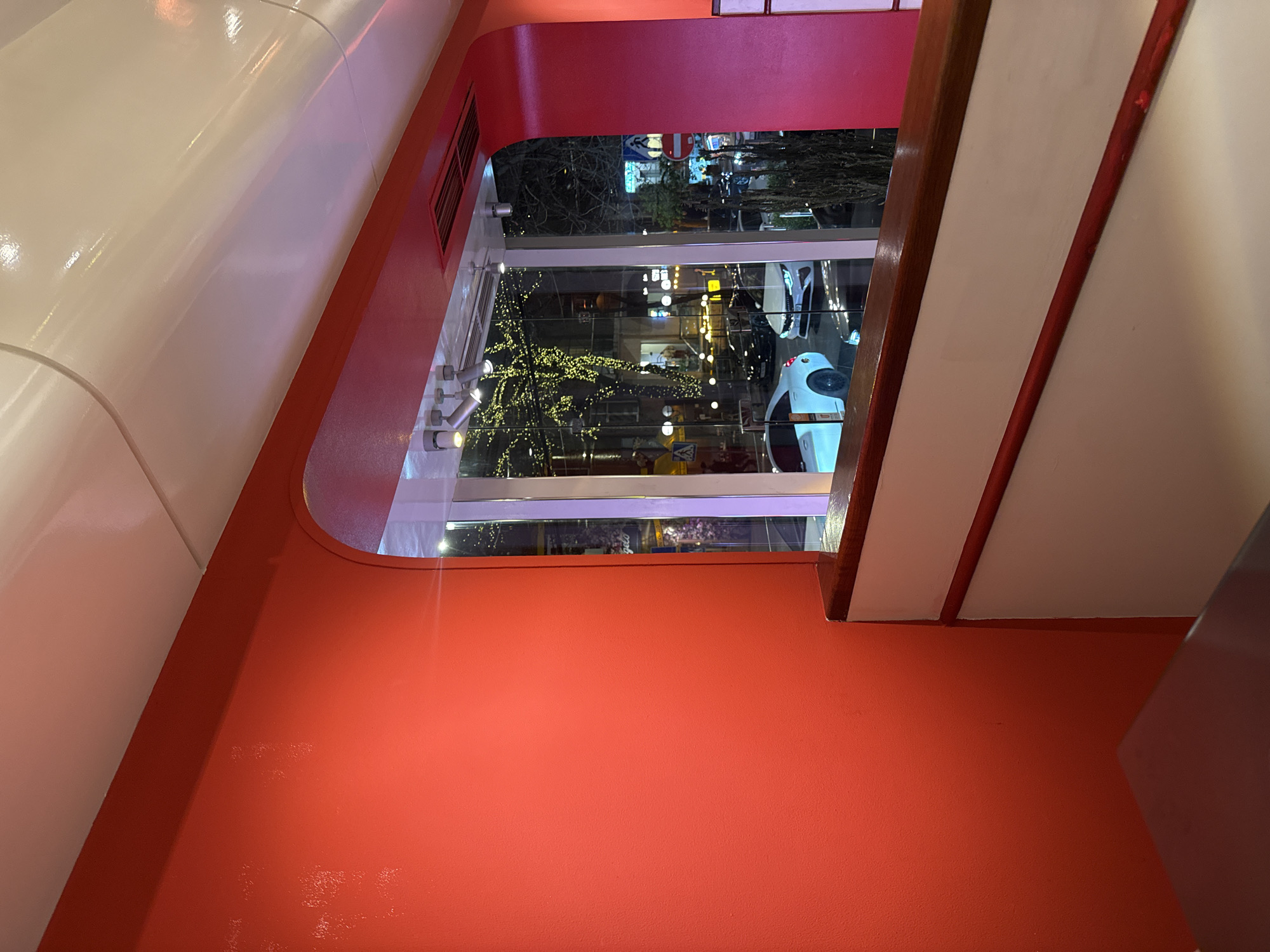 The image depicts a street view through a window from an elevated interior location. The interior features red walls and white trim, creating a geometric frame around the window. The window overlooks a street in Yerevan, Armenia, showing traffic and pedestrians at night. The street is lit with streetlights, illuminating trees decorated with lights. Vehicles, including white cars, are visible on the road. The angle of the shot suggests it was taken from inside a building, possibly a restaurant or cafe, looking down onto the street.