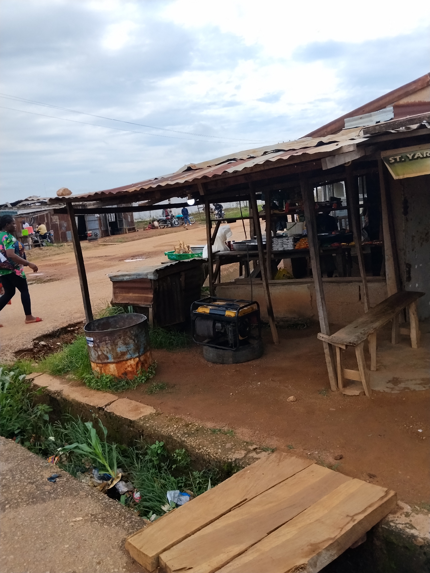 This image captures a street scene in Mangu Town, Nigeria, likely a market area. In the foreground, a woman in a colorful top and black leggings walks on a dirt path, her red sandals visible. To her left, a rusty metal barrel stands amidst green vegetation and a drainage ditch filled with debris. A generator is situated on a tire near the barrel. The main subject is a small outdoor shop or stall with a corrugated metal roof supported by wooden poles. Inside, shelves are stocked with goods, and a person can be seen behind the counter. A wooden bench is placed to the right of the stall. In the background, other dilapidated structures, some with corrugated metal roofs, line the dirt road. Several motorcycles are parked, and people are visible in the distance, suggesting ongoing activity. The sky is overcast with clouds. The overall impression is one of a developing urban environment with informal commercial activities.