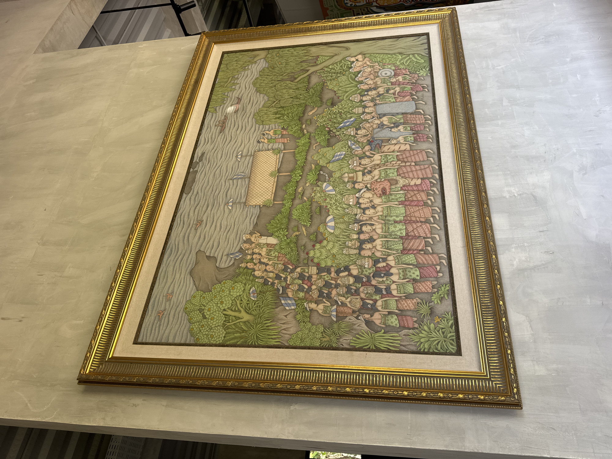 This image shows a large, rectangular painting framed in ornate gold, hung on a rough, gray concrete wall. The painting depicts a procession of many people, seemingly participating in a ceremony or parade. They are dressed in traditional attire, primarily sarongs in shades of green, pink, and red, with some wearing head coverings. Many individuals carry offerings or baskets on their heads.
The scene is set in an outdoor environment with lush greenery, trees, and a body of water in the background. There are also some structures that appear to be sheds or temporary shelters, along with umbrellas scattered among the crowd. Small boats are visible on the water, and birds are depicted in the sky and near the water. The painting's style is detailed and somewhat stylized, with vibrant colors used to illustrate the figures and the natural surroundings. The overall impression is one of a community event or cultural ritual taking place in a tropical setting.
The photograph was taken in Kuta Selatan, Indonesia, suggesting the painting likely represents a Balinese cultural scene.