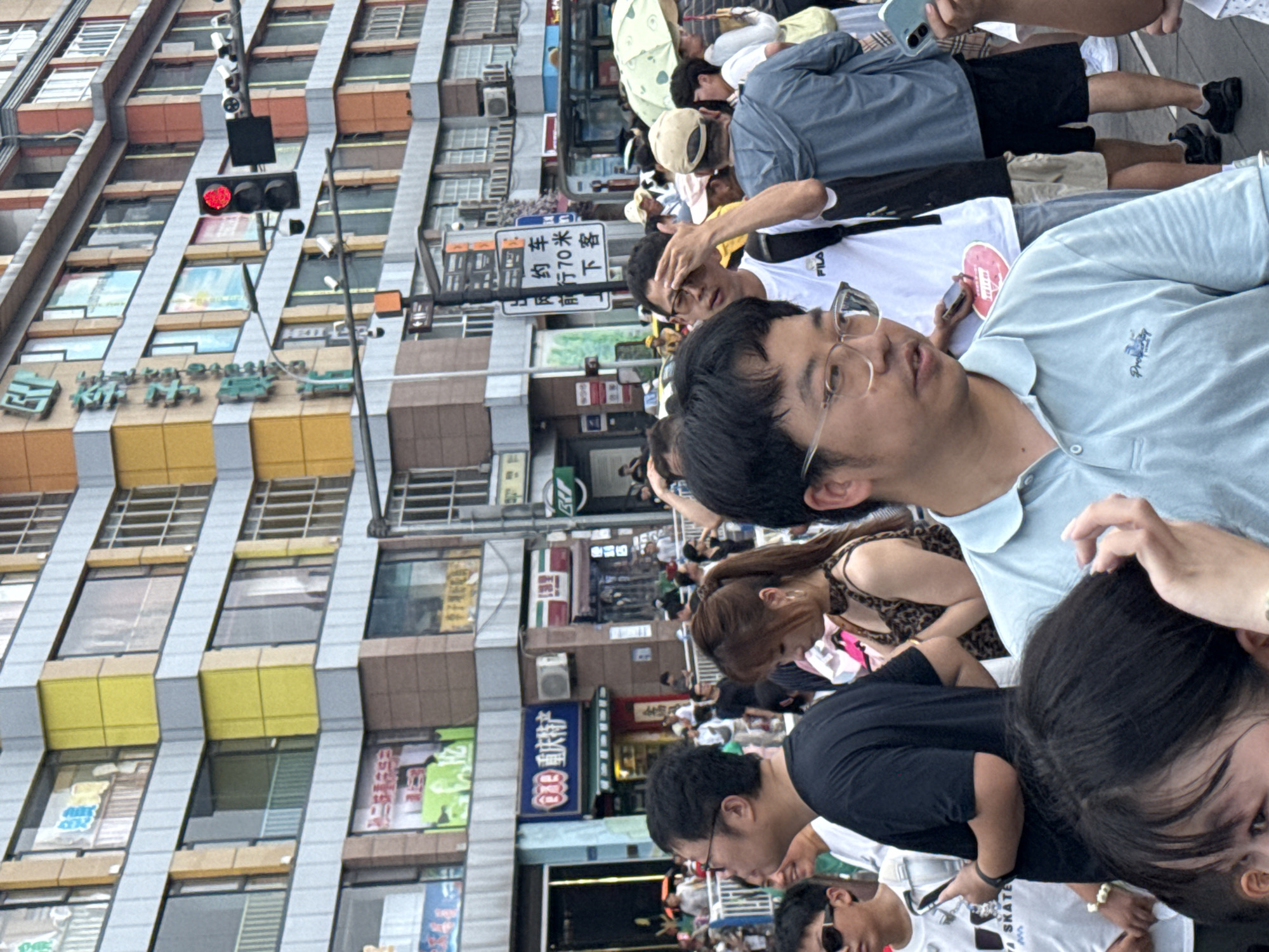 This is a street-level, medium-distance photograph taken in the Yuzhong District of China. The scene depicts a bustling urban environment with a crowd of people in the foreground and midground, a multi-story building with shops on the ground floor in the background, and a traffic light displaying red. The building features a colorful facade with yellow, orange, and grey panels, and numerous windows. Several signs with Chinese characters are visible on the building's exterior.
In the foreground, a man wearing glasses and a light blue polo shirt is looking to the right. To his left, partially visible, is a young person wearing sunglasses and a white t-shirt. Behind the man in the blue shirt, other individuals are present, including a woman with long brown hair wearing a leopard-print top.
The midground shows a dense gathering of people navigating a pedestrian area. Some individuals are holding umbrellas, suggesting sunny weather. A bus and other vehicles are partially visible further down the street.
The overall atmosphere appears to be one of active commerce and public transit, typical of a commercial district in a Chinese city. The red traffic light indicates that vehicles are stopped, and the crowd suggests a busy time of day or a special event.
