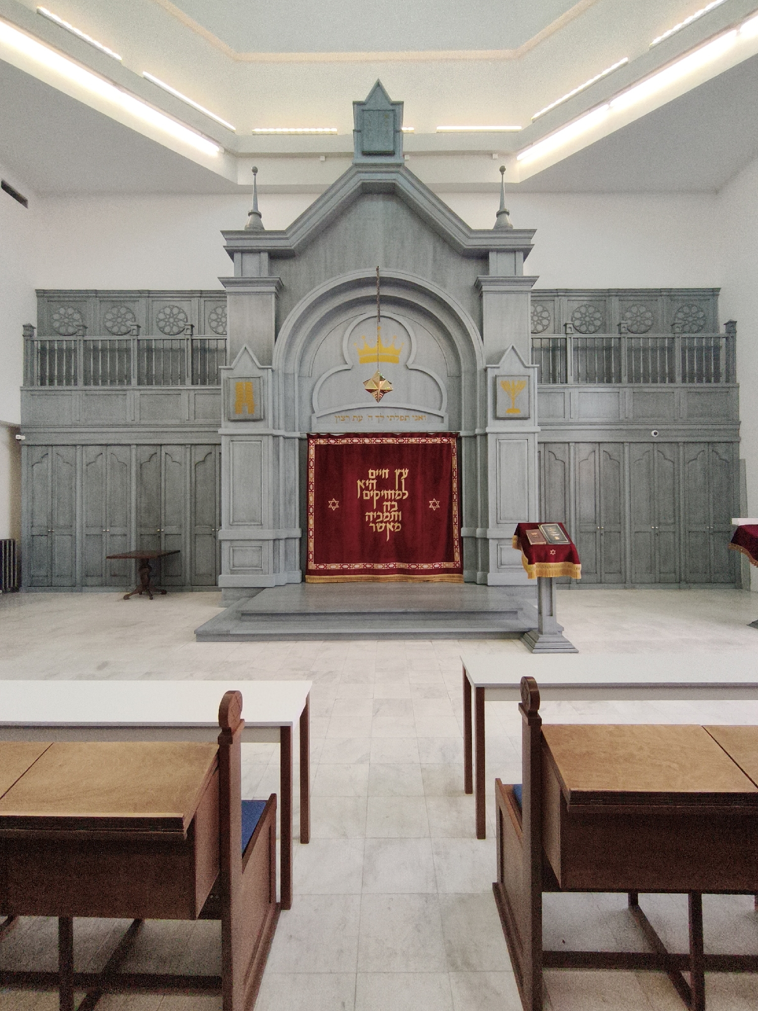 The image depicts the interior of a synagogue. The focal point is a grand ark with a red curtain, featuring Hebrew inscriptions and a Star of David. To the right of the ark, a wooden lectern holds two open books covered by a gold-fringed cloth. The synagogue's architecture includes ornate grey wooden panels, railings, and a tiered structure above the ark. In the foreground, two rows of long, light-colored tables are visible, with two wooden benches with blue cushions placed in front of them. The floor is tiled with a light-colored marble pattern. This synagogue is located at 3a Chernyakhovsky Street in Kaliningrad, Russia. There are no people in the image.