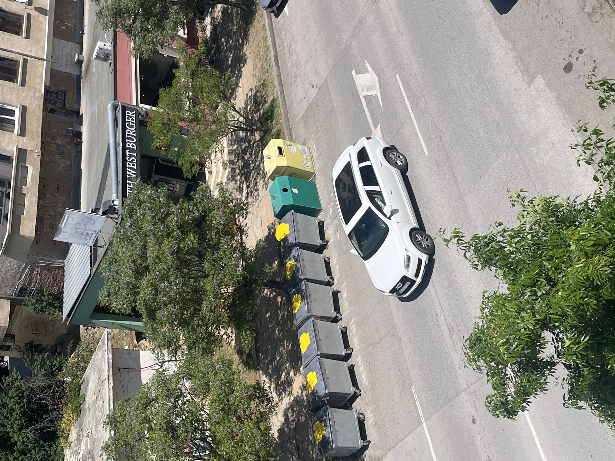 The image shows an aerial view of a street in Blagoevgrad, Bulgaria. A white car is parked on the right side of the street next to a row of gray and yellow waste bins. A yellow and a green recycling bins stand at the beginning of the row of gray bins. A white arrow is painted on the street, indicating the direction of traffic. On the left side of the street is a building with a sign that reads "SOUTH WEST BURGER". Trees line both sides of the street. The weather appears to be sunny.