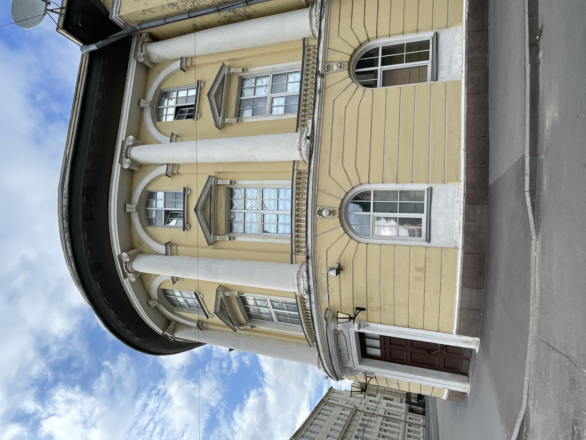The image captures a portion of the curved facade of a yellow building with neoclassical architectural elements in Saint Petersburg, Russia. The building features large, white Corinthian columns supporting an entablature. Several windows are visible, some rectangular with white frames and grid-like panes, and others are arched. A dark wooden door with a rectangular window is set into an arched entryway. Wall-mounted lanterns are positioned on either side of the door. The building is bordered by a grey sidewalk and an asphalt road. In the background, a neighboring building with numerous windows can be seen. The sky is partly cloudy with scattered white clouds against a blue background. The perspective is from a low angle, looking upwards at the building.