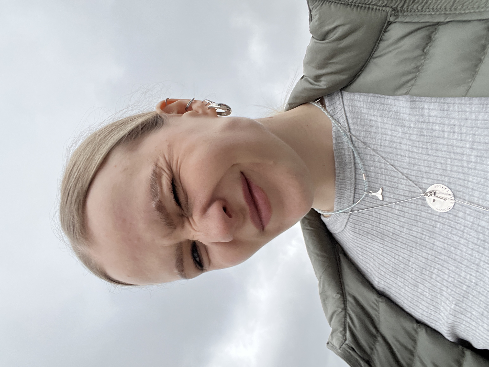 A woman with fair skin and blonde hair pulled back is depicted from a low angle, looking down at the camera with a squinted expression. She is wearing multiple silver earrings on her left ear and two silver necklaces. She is wearing a grey ribbed top and a padded olive-green vest. The background is an overcast sky. There is no visible evidence confirming the city of Khimki, Russia.