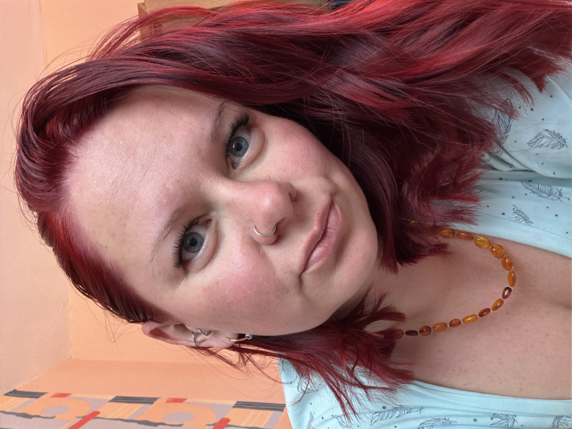 An adult female is depicted in a close-up, head-and-shoulders portrait, situated indoors in the city of Chaikovsky, Russia. She possesses dark red, shoulder-length hair showing darker roots, and blue eyes that meet the camera's gaze directly. Her skin is fair, with visible freckles on her face and upper chest. A silver hoop pierces her left nostril. Her visible left ear (viewer's right) features multiple piercings, including a silver hoop in the helix and a small gold-colored hoop with a bead in the lobe. She wears a light blue top patterned with white feathers and an amber-colored beaded necklace around her neck. The background consists primarily of a plain light orange wall, with a section of geometric wallpaper or artwork featuring orange, grey, and white abstract shapes visible behind her left shoulder. The scene is illuminated by soft, even lighting.