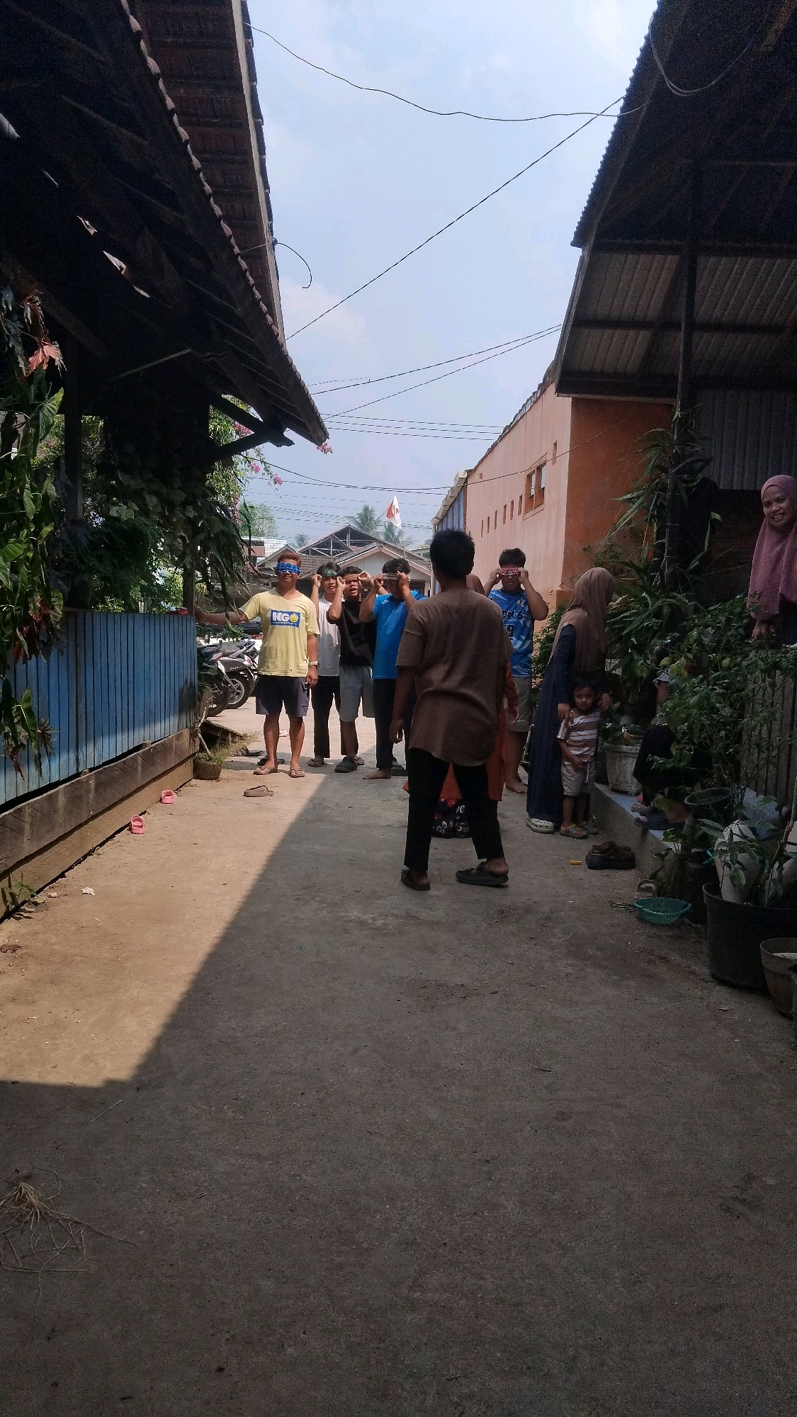 The image depicts an outdoor scene in what appears to be a residential alleyway in Bangun Rejo, Indonesia. A group of young men, all appearing to be of Indonesian descent, are engaged in an activity, possibly a game. Several of them have blue cloths or headbands tied around their eyes, suggesting a blindfolded game is in progress. The alley is paved with concrete and flanked by buildings on both sides. On the left, there is a low blue wooden fence partially obscured by dense green foliage. On the right, an orange building with visible windows stands alongside a narrow path lined with potted plants and a few individuals, including two women in headscarves and a small child. In the background, several motorcycles are parked. The sky is clear and bright, indicating daytime. The overall atmosphere suggests a casual, community gathering or recreational activity.