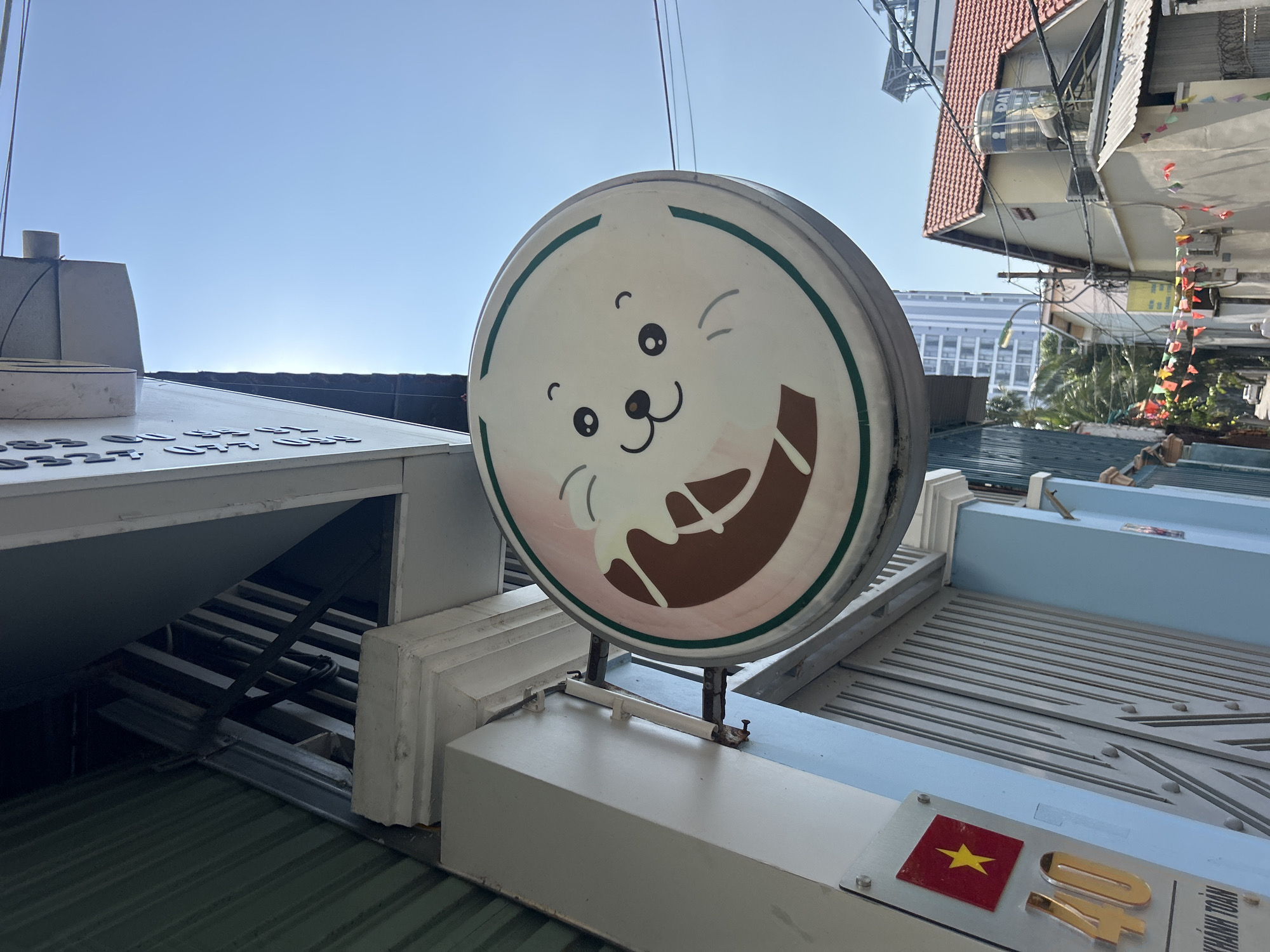 The image shows a round sign with a cartoon seal face on it. The sign is white with a green border and the seal face has black eyes, nose, and whiskers. There is a brown portion on the bottom right of the seal image that resembles a melted chocolate drink or ice cream. The sign is mounted on a platform on top of a building, possibly a restaurant or cafe. The platform is painted white and blue. There is a Vietnamese flag and some golden letters. The environment appears to be a building rooftop in an urban area. From the sign plate in the image, the location is inferred to be in the city of Bac Nha Trang Ward, Vietnam. The sky is clear and blue. Other buildings are visible in the background.