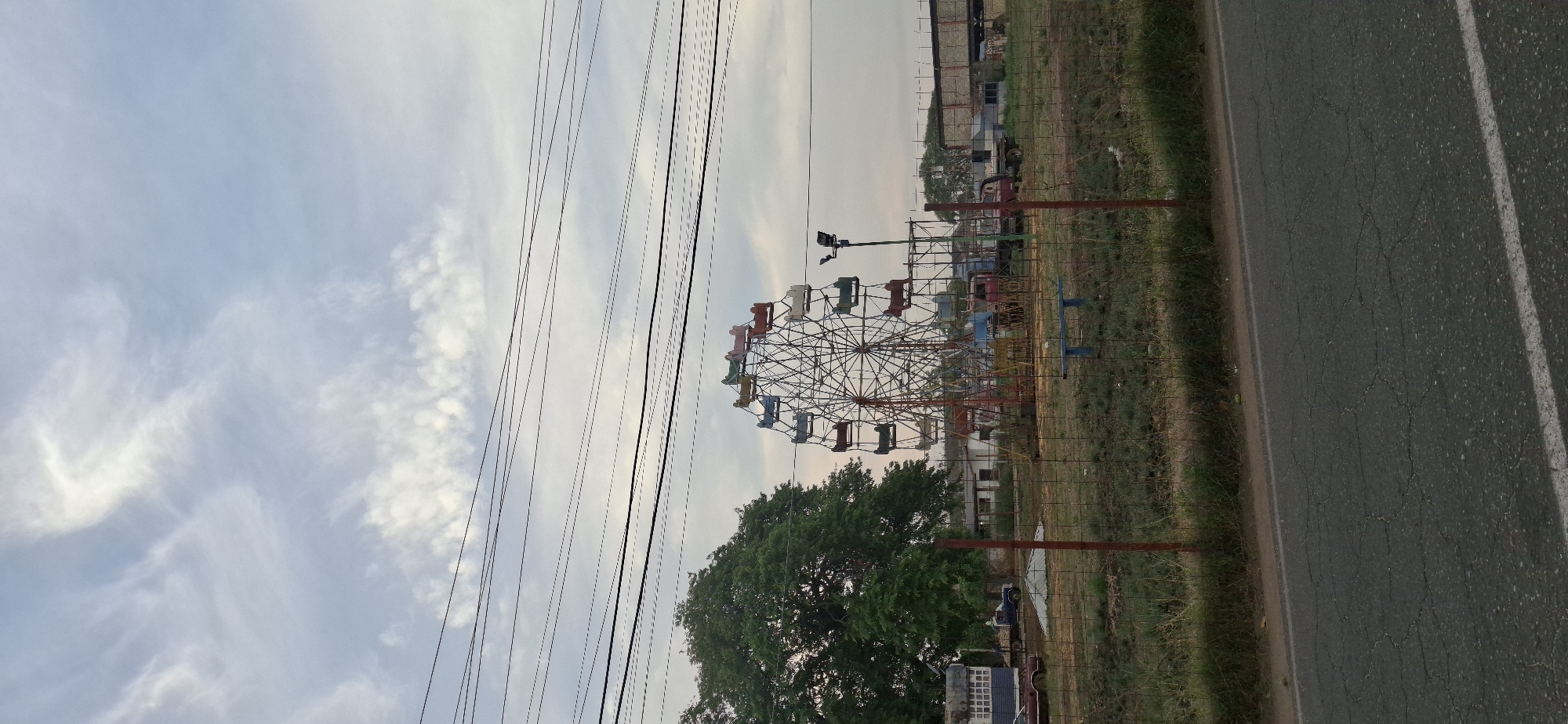 The image displays an outdoor scene in Tinaquillo, Venezuela, centered around a stationary Ferris wheel. The Ferris wheel is a metal structure featuring multiple cabins in various colors including red, orange, green, blue, white, and brown. It stands in a grassy area, with a large, lush green tree partially obscuring its lower section.
Numerous utility lines and poles crisscross the sky in the upper left and central parts of the frame. A wire mesh fence with reddish-brown posts separates the Ferris wheel area from a cracked, dark asphalt road with a white painted line, visible on the right side of the image. In the background, beyond the Ferris wheel, several indistinct buildings and structures can be seen, along with what appear to be parked vehicles or other equipment. A blue metal bench is also visible near the Ferris wheel.
The sky is a pale blue with scattered, wispy white clouds, suggesting either early morning or late afternoon lighting. No people or active events are captured in the scene, which conveys a sense of quietude. There is no visible text.