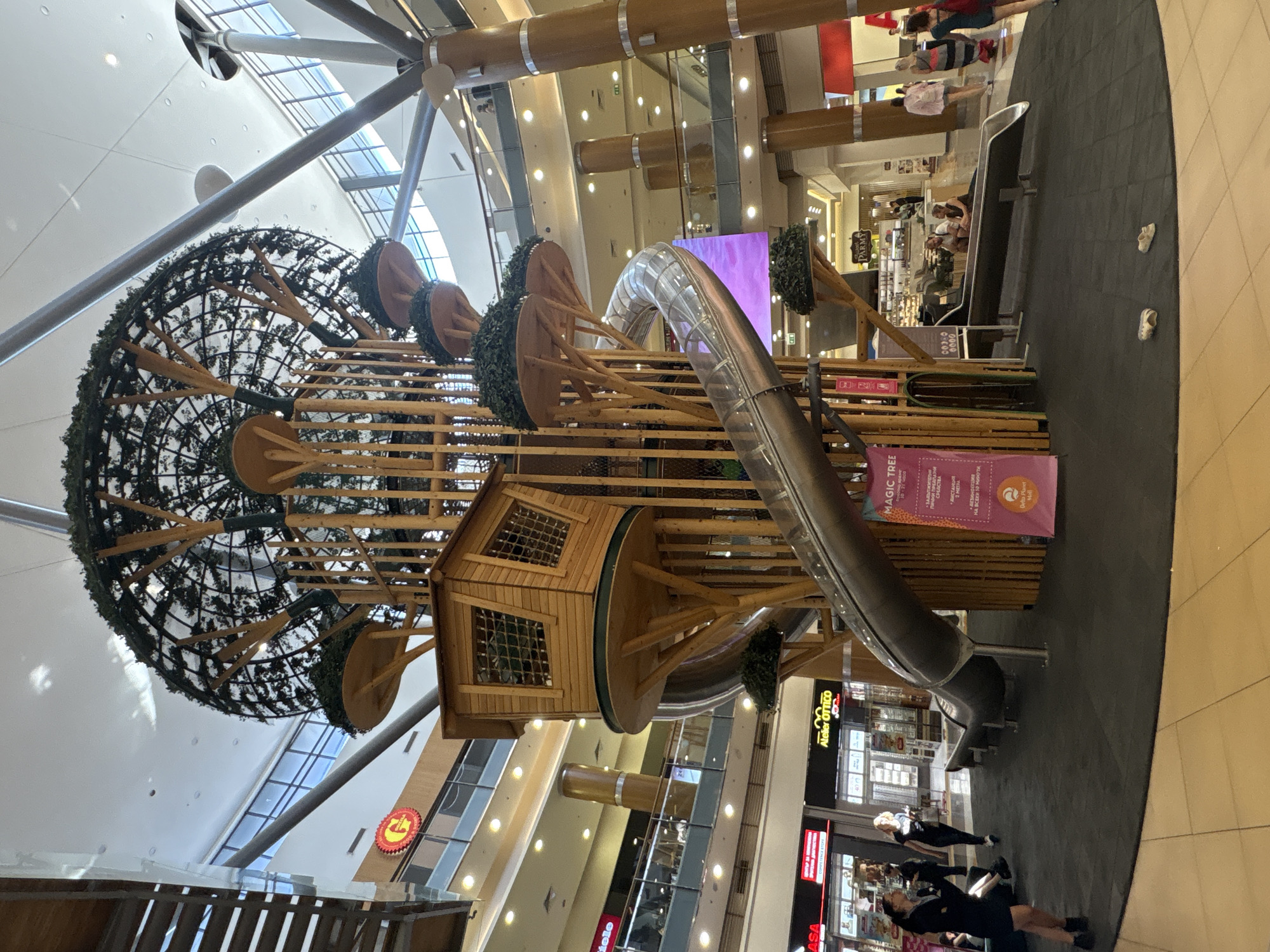 This is a vertically oriented, full-shot photograph taken inside a modern shopping mall, likely during daylight hours given the natural light filtering through the ceiling. The main subject is a large, elaborate indoor playground structure designed to resemble a treehouse or a forest.
The playground features a central wooden structure with a small house-like section with windows. Extending from this are branches made of wood, adorned with large, circular platforms covered in green foliage, giving the impression of leaves or tree canopies. A large, curved metal slide is integrated into the structure, swooping downwards from the upper levels. The entire playground is constructed with natural-looking materials like wood and metal, with a black padded floor surrounding its base.
In the background, the multi-level interior of the mall is visible, with shops, escalators, and retail signage. The architecture suggests a contemporary commercial space with large windows and exposed structural elements. A few people are scattered throughout the mall, some appearing to be waiting or browsing near the shops. There are also a few pairs of children's shoes left on the black matting near the slide, suggesting the playground is actively in use by children.
Visible text includes "MAGIC TREE", "Atelier OTTICO", and "PARMY" on shop signs and a banner on the playground structure. The banner also contains Cyrillic text, indicating the location is likely in a region where this script is used, possibly Eastern Europe. The overall atmosphere appears to be lively and family-friendly, typical of a shopping mall entertainment area.